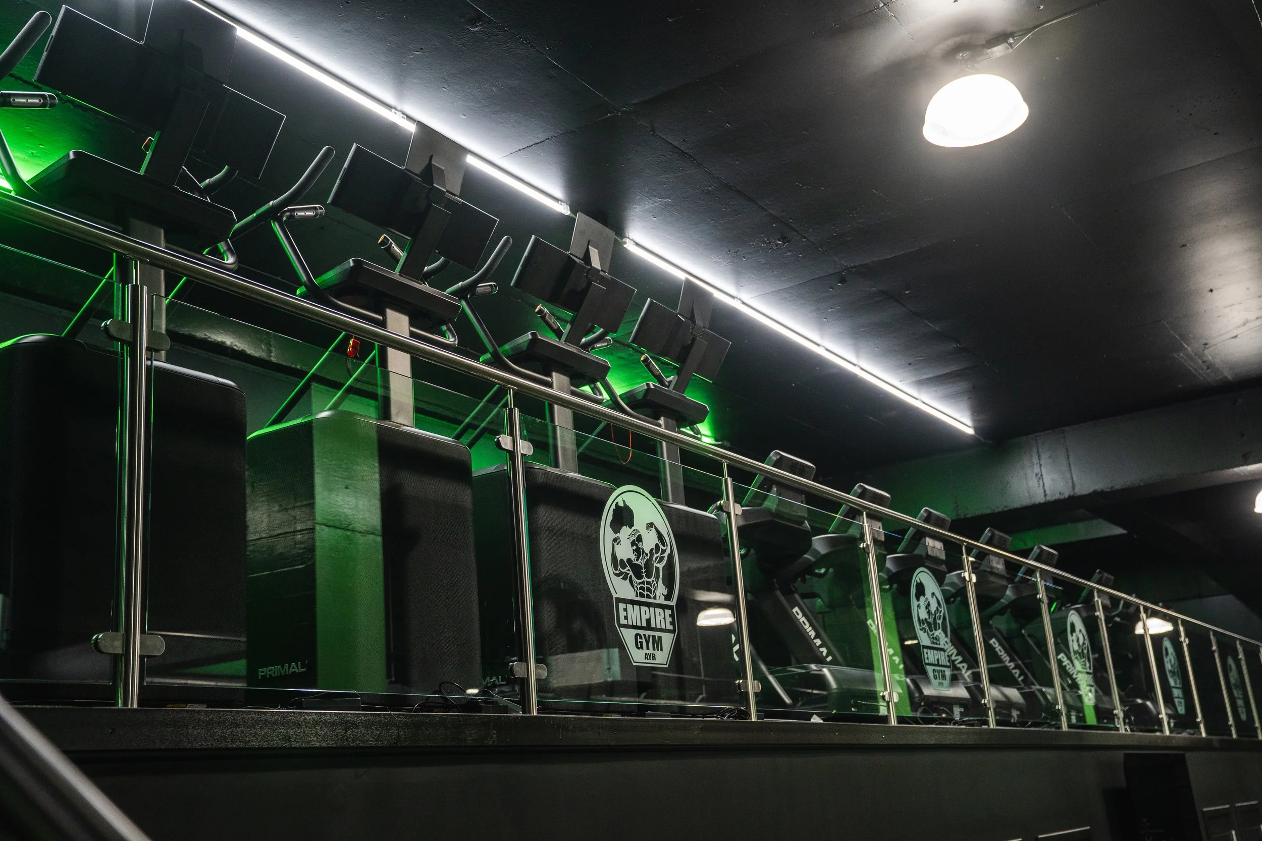 Row of black workout machines with the Empire Gym logo on a sign, illuminated by green lights, inside a gym with dark walls and ceiling.