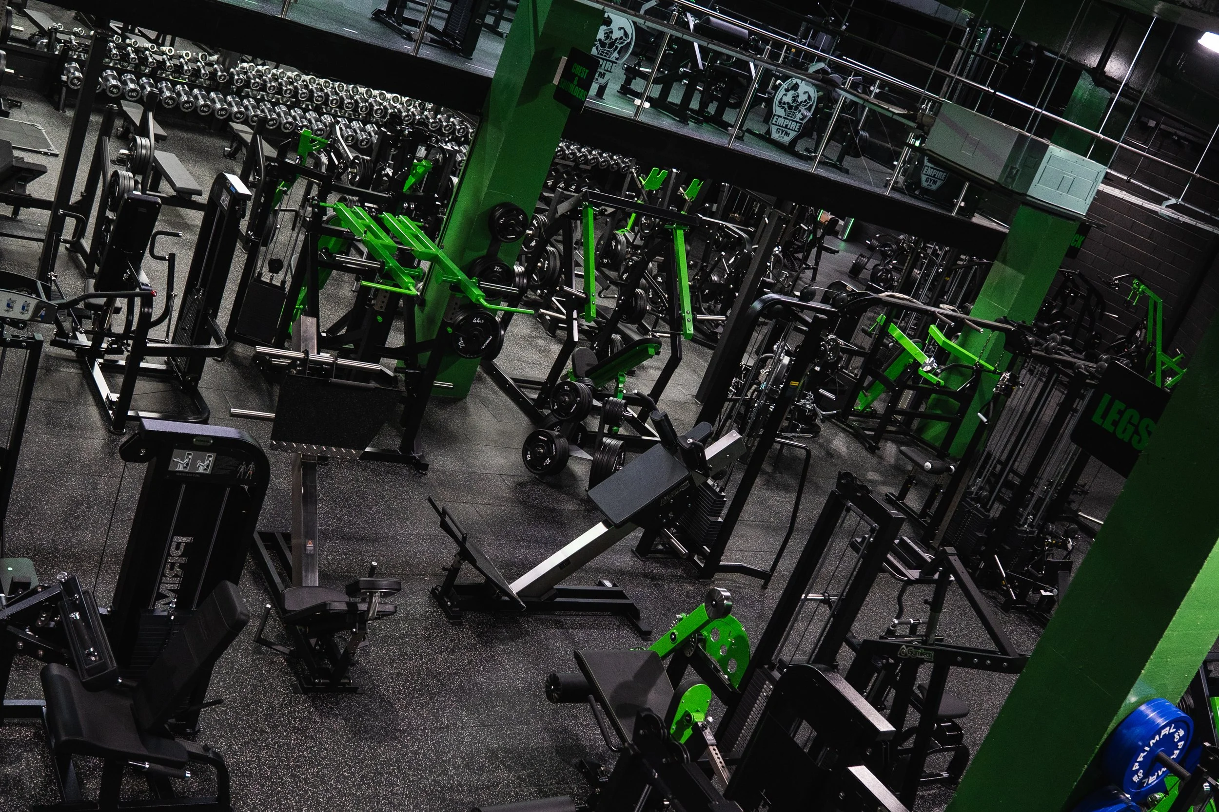 Interior of a gym with various black and green weightlifting equipment, including dumbbells, benches, and machines, with mirrors and black flooring.