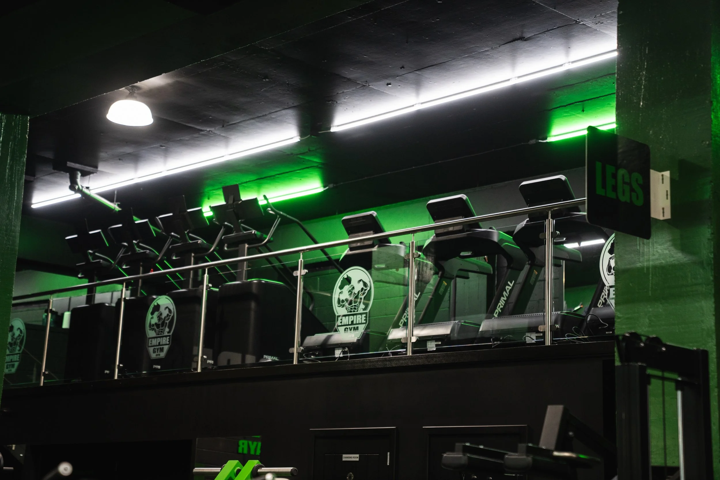 Gym equipment on a second floor balcony, including treadmills with Empire Gym logos and a glass railing, illuminated by bright white and green lighting.