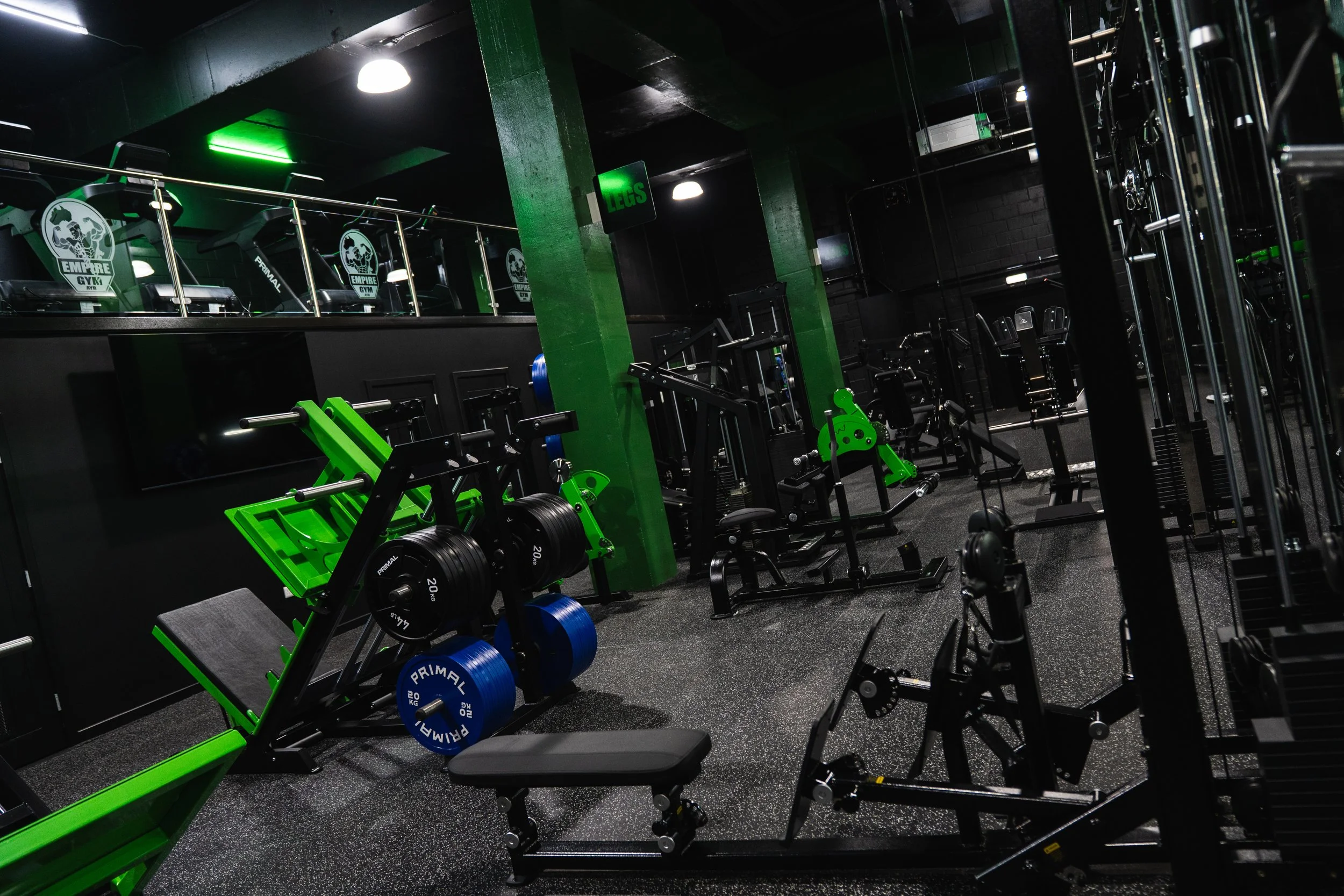 A gym with various workout machines and free weights, including a green and black weightlifting bench, and dumbbells with weights, in a dark room with black walls and bright lighting.