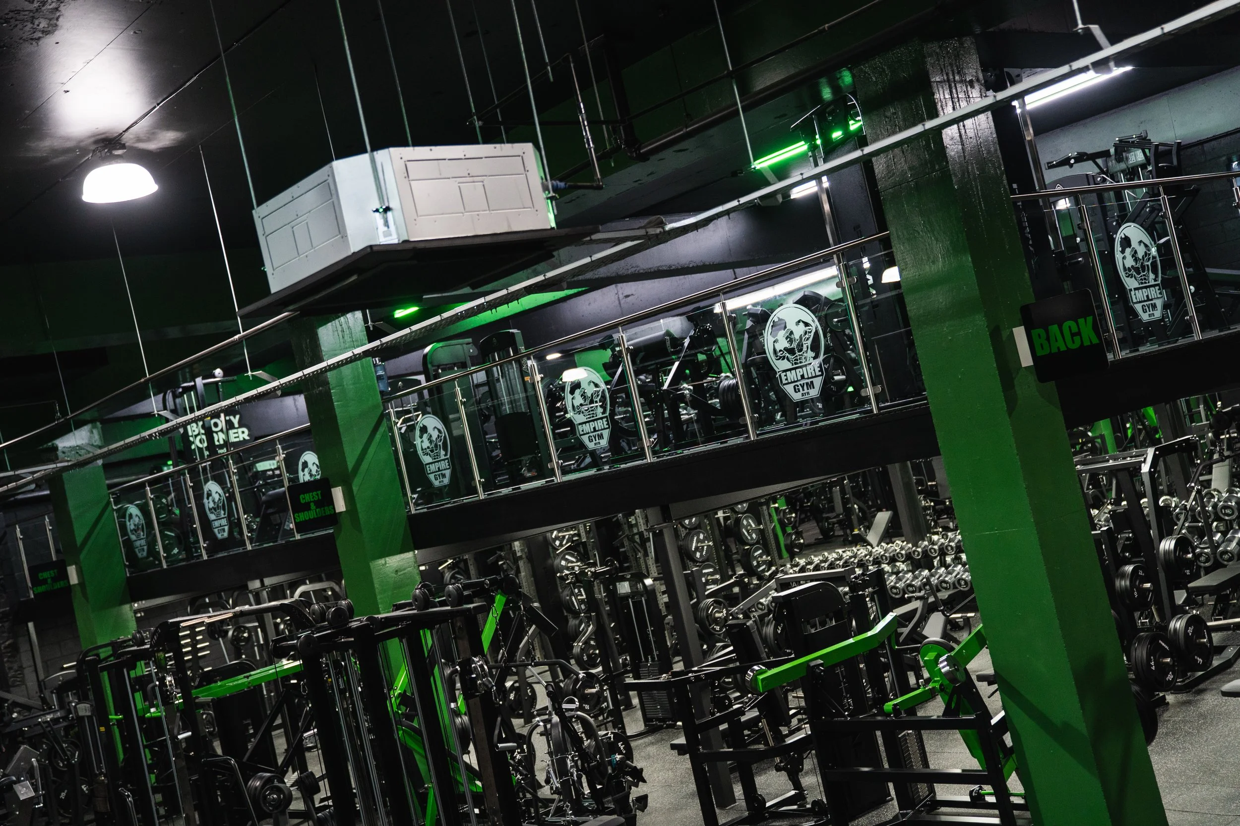 Interior of a gym with rows of weightlifting equipment and mirror walls, featuring black and green color scheme, with signs labeled 'CHEST & SHOULDERS' and 'BACK'.