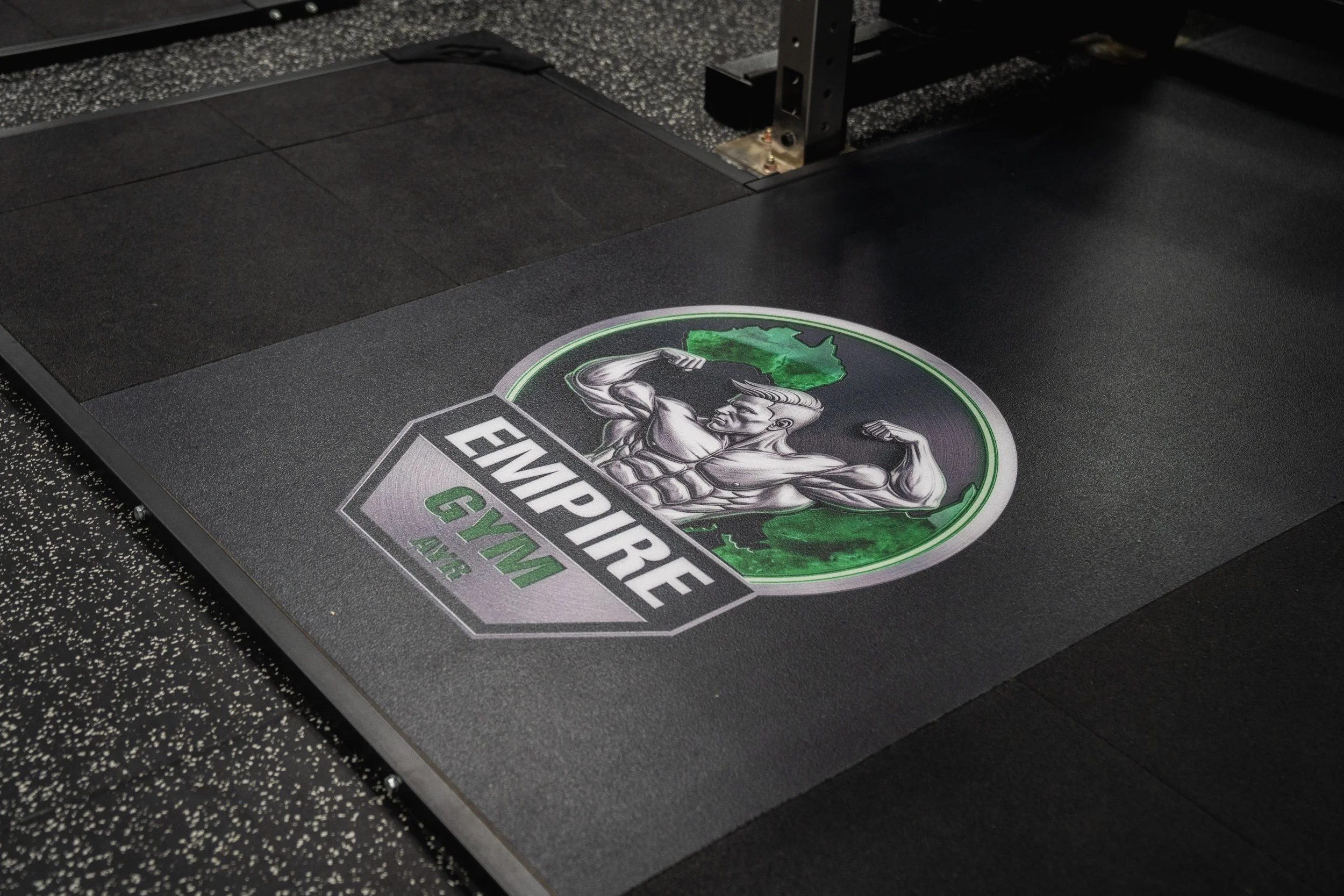 Close-up of a black exercise step platform with an logo of a muscular cartoon figure flexing, accompanied by the text 'Everpure Gym'. The platform is placed on a speckled black and gray floor, and part of a metallic frame is visible.