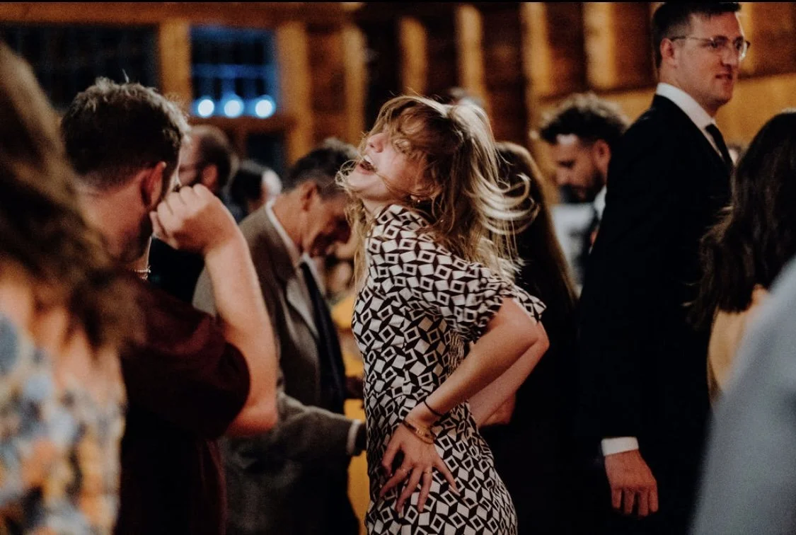 Woman with curly hair dancing and smiling at a social gathering in a wooden indoor venue.