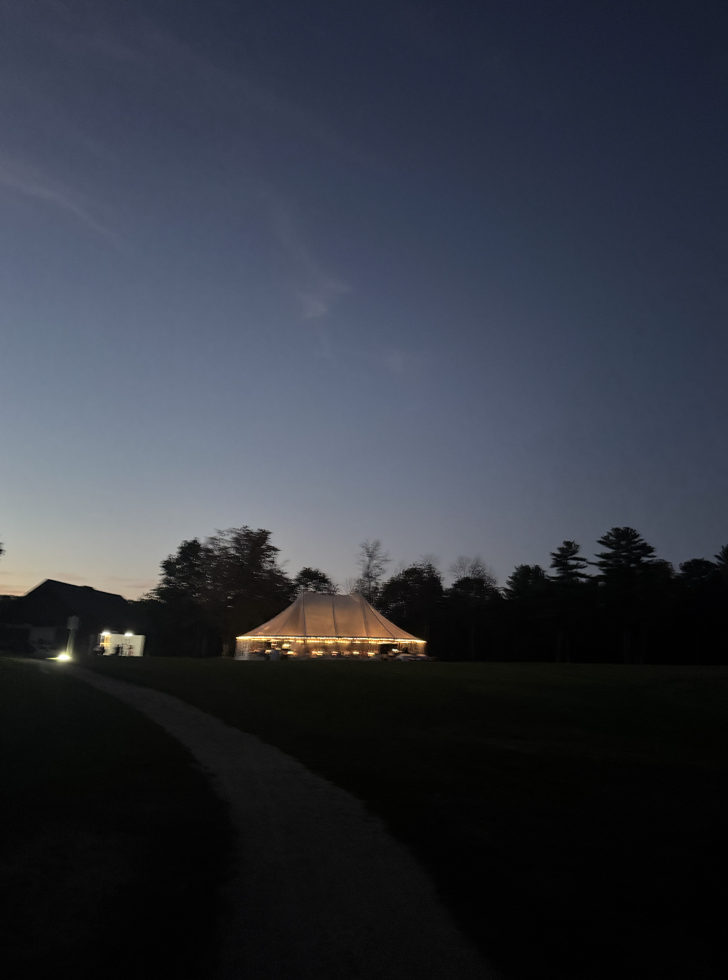 A large illuminated tent with string lights at dusk, surrounded by trees and a dark sky.
