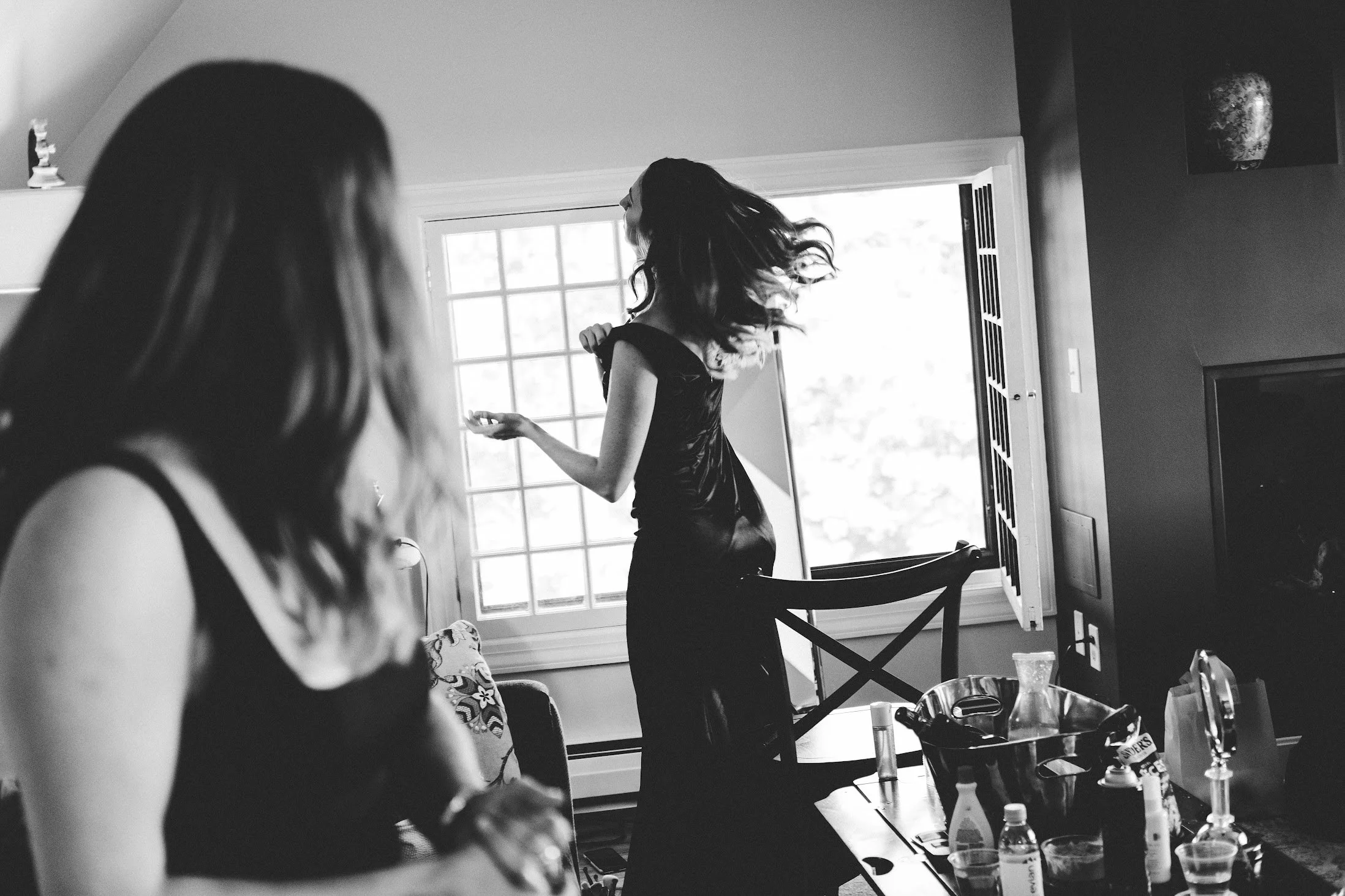 A woman with long dark hair dancing in a room with a large window, while another woman with shoulder-length hair, wearing a black tank top, is sitting near a table with various bottles and cups.