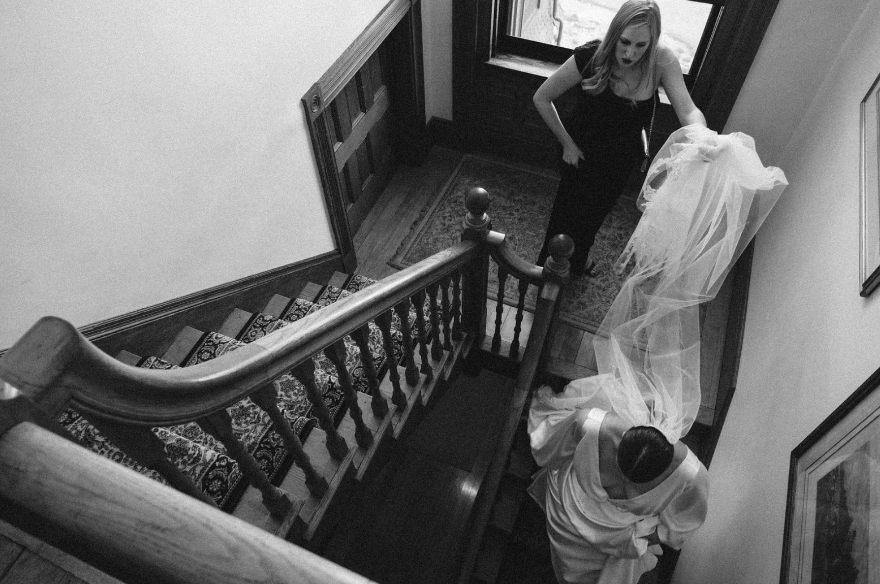 A woman in a black dress is standing at the top of a staircase, holding a long, flowing piece of sheer fabric, with another person assisting her, and a man at a lower level looking up. The scene is captured from a high angle in black and white.