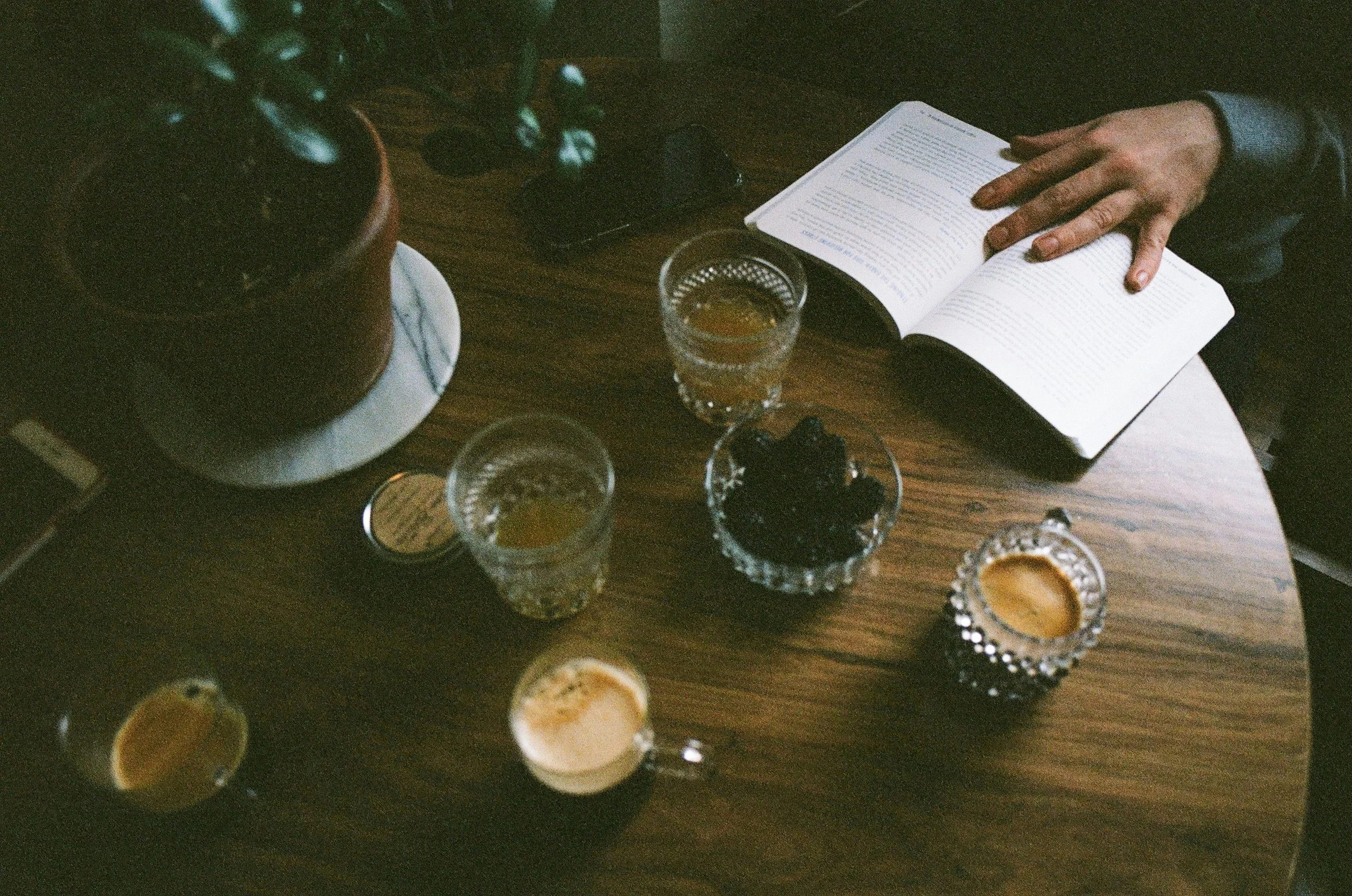 A person with long nails reading a book at a wooden table with several shot glasses filled with different beverages, some with orange-colored liquid and others with dark-colored liquid, and a potted plant.