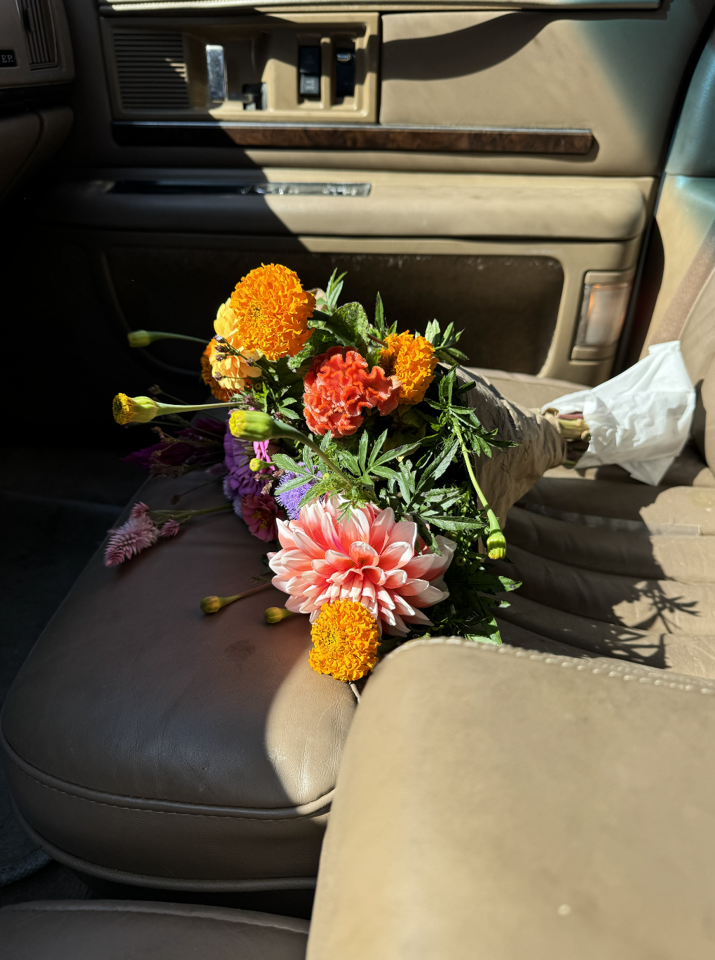A bouquet of colorful flowers, including marigolds, a pink dahlia, and other blossoms, resting on a beige car seat with tissue paper wrapped around the stems.