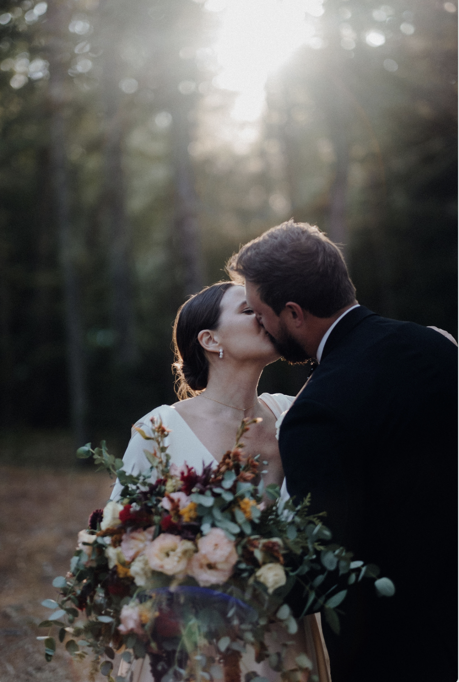 A bride and groom share a kiss outdoors during sunset, with the bride holding a large bouquet of flowers and wearing a white dress, and the groom in a dark suit, surrounded by trees.