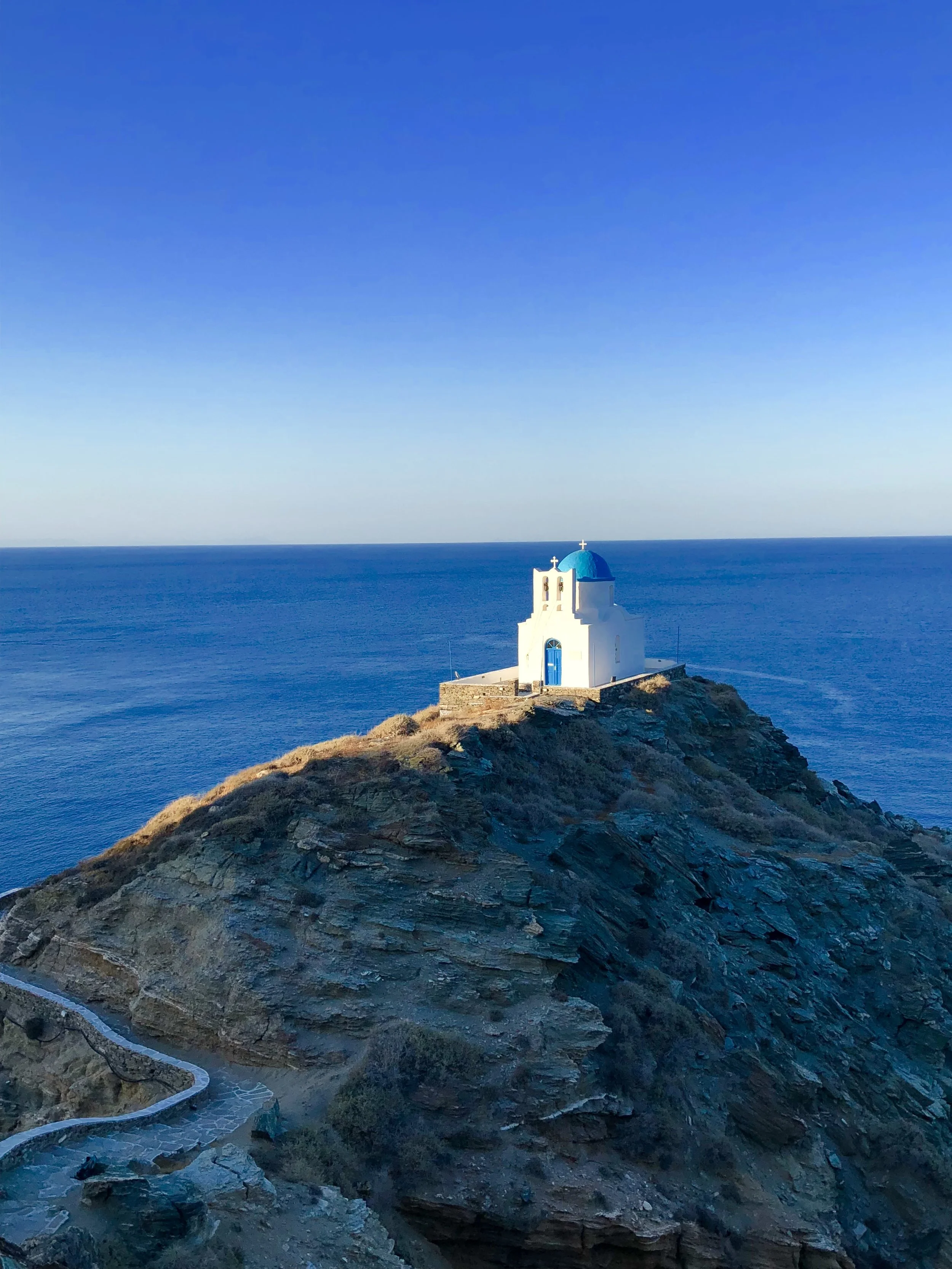 A white church with a blue dome and cross on top, situated on a rocky hillside overlooking the ocean under a clear blue sky.