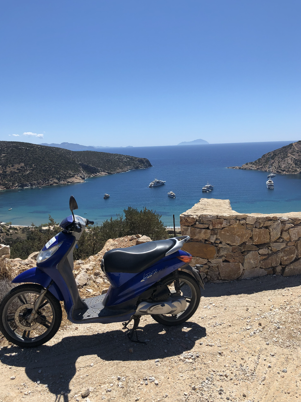 A blue scooter parked on a dirt path overlooking a bay with several boats and yachts anchored in the water, with hills and islands in the background under a clear blue sky.
