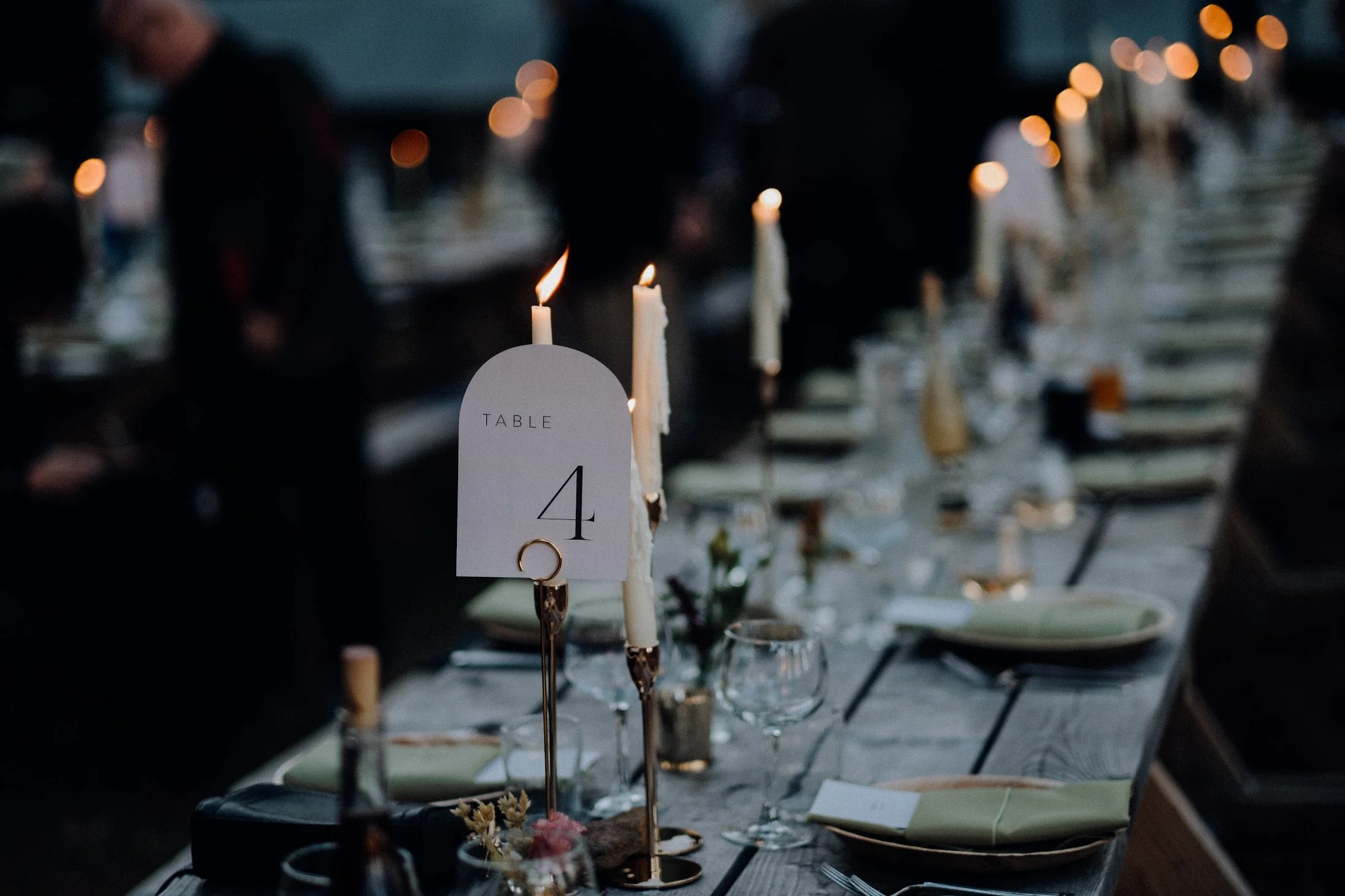A blurred image of a dinner table set for an event, decorated with candles, flowers, and place settings, with a prominent table number sign indicating table 4, and guests in formal attire in the background.