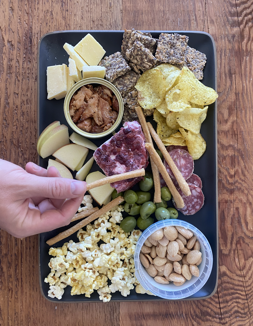 Charcuterie plate with cheese, crackers, fruit, popcorn, almonds, canned dip, potato chips, beef jerky, sausage, green olives, and a hand reaching for crackers.