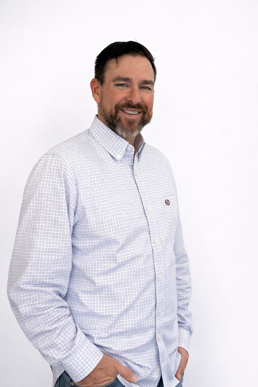 A man with dark hair and a beard smiling, wearing a light-colored checkered button-up shirt with his hands in his pockets, standing against a plain white background.