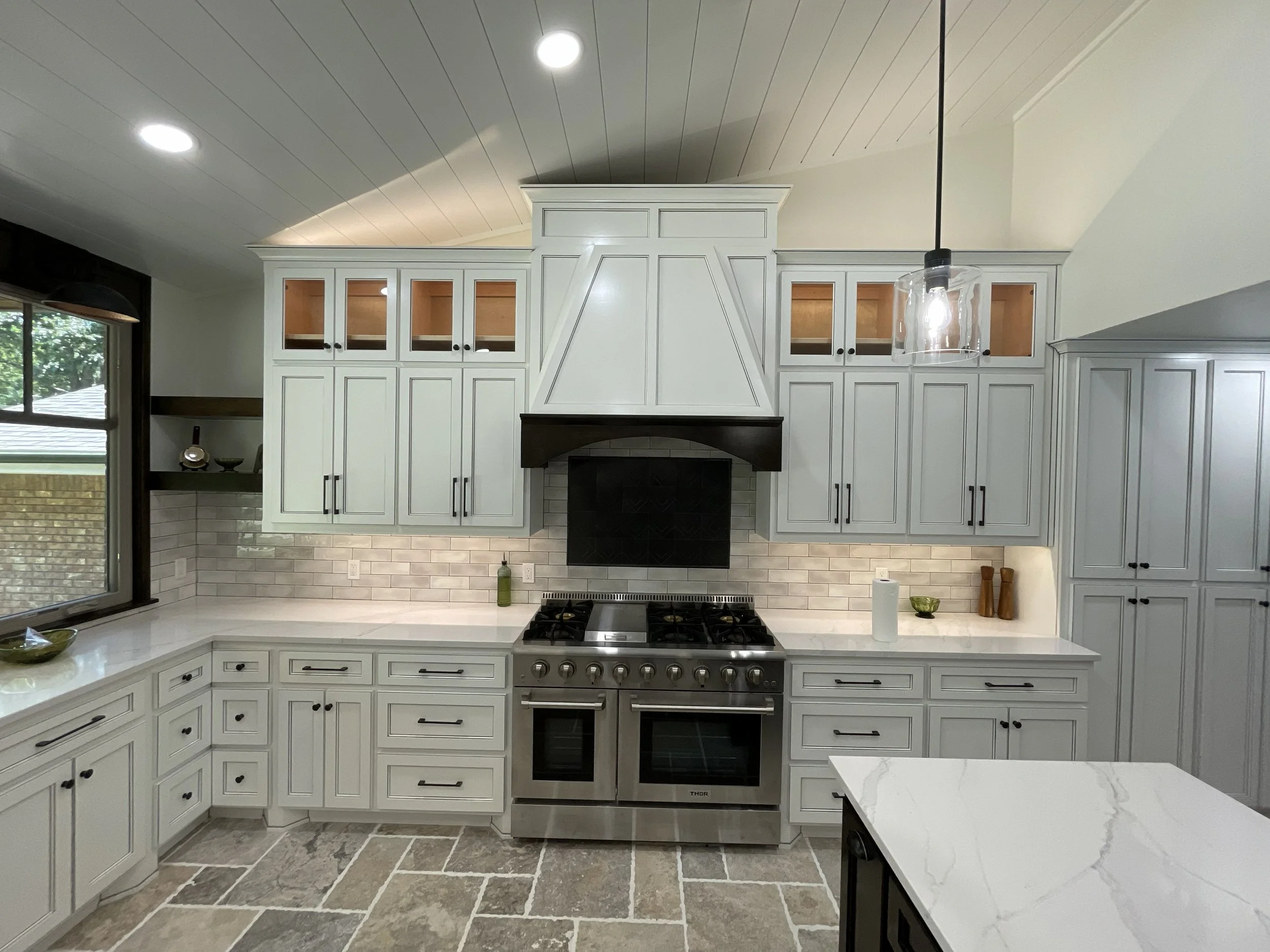 Kitchen with white cabinets, stainless steel oven, and a stone tile floor. There are large windows on the left with a view of trees outside. The ceiling has recessed lighting, and a pendant light hangs above the countertop on the right.