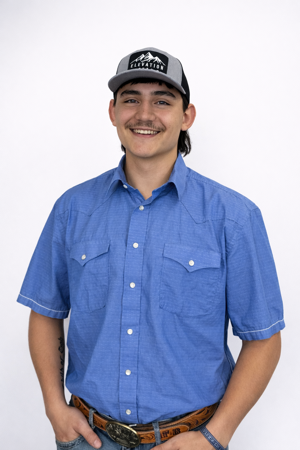 Smiling young man wearing a blue button-up shirt, gray baseball cap with mountains and 'Elevation' logo, posing against a plain white background.