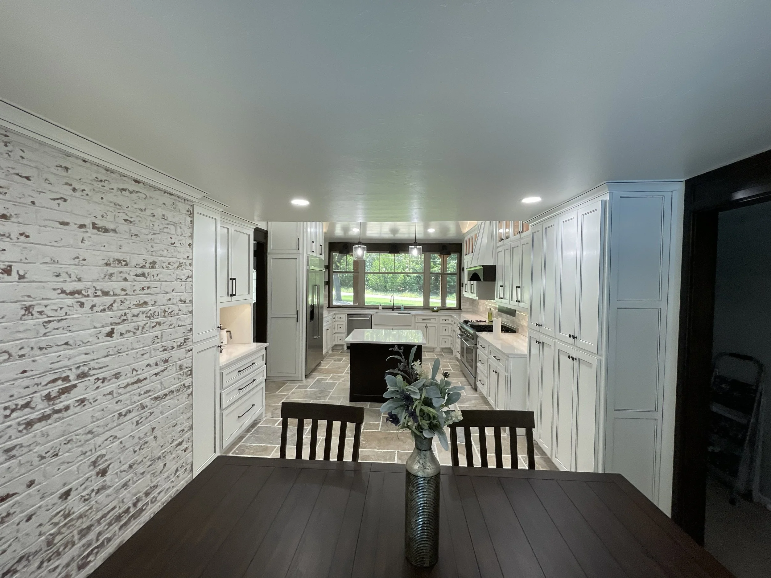 View of a bright, spacious kitchen with white cabinets, black accents, large windows, and gray tiled flooring, with a dining table and chairs in the foreground.