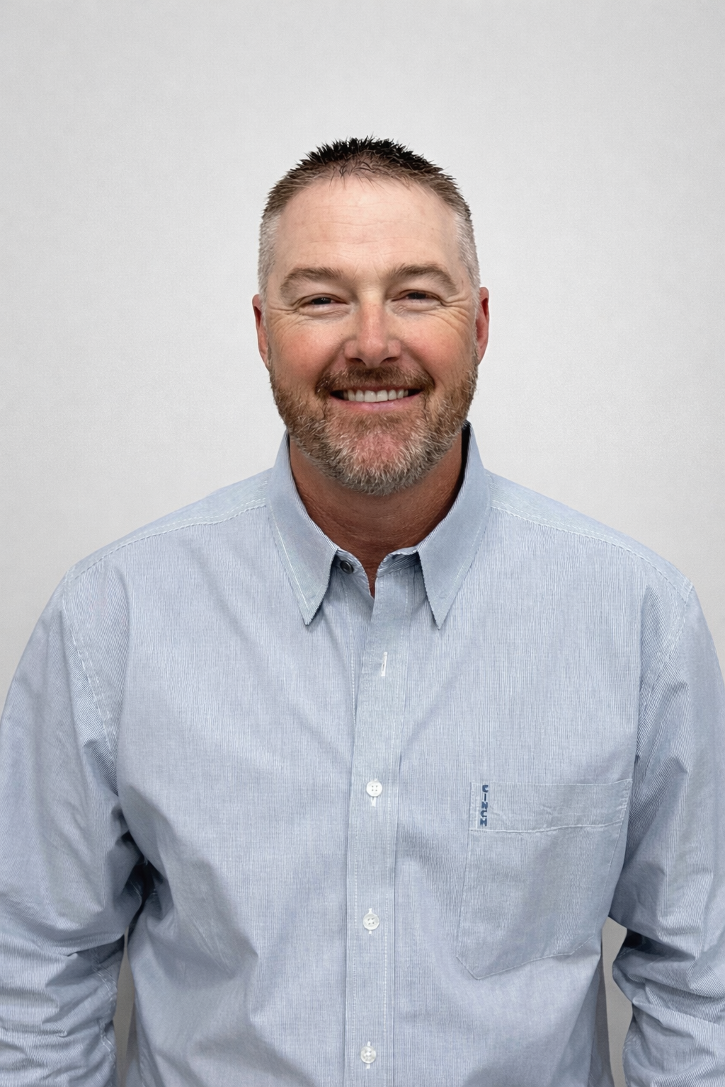 A smiling middle-aged man with short light brown hair, a beard, and blue eyes, wearing a light blue button-up shirt against a plain white background.
