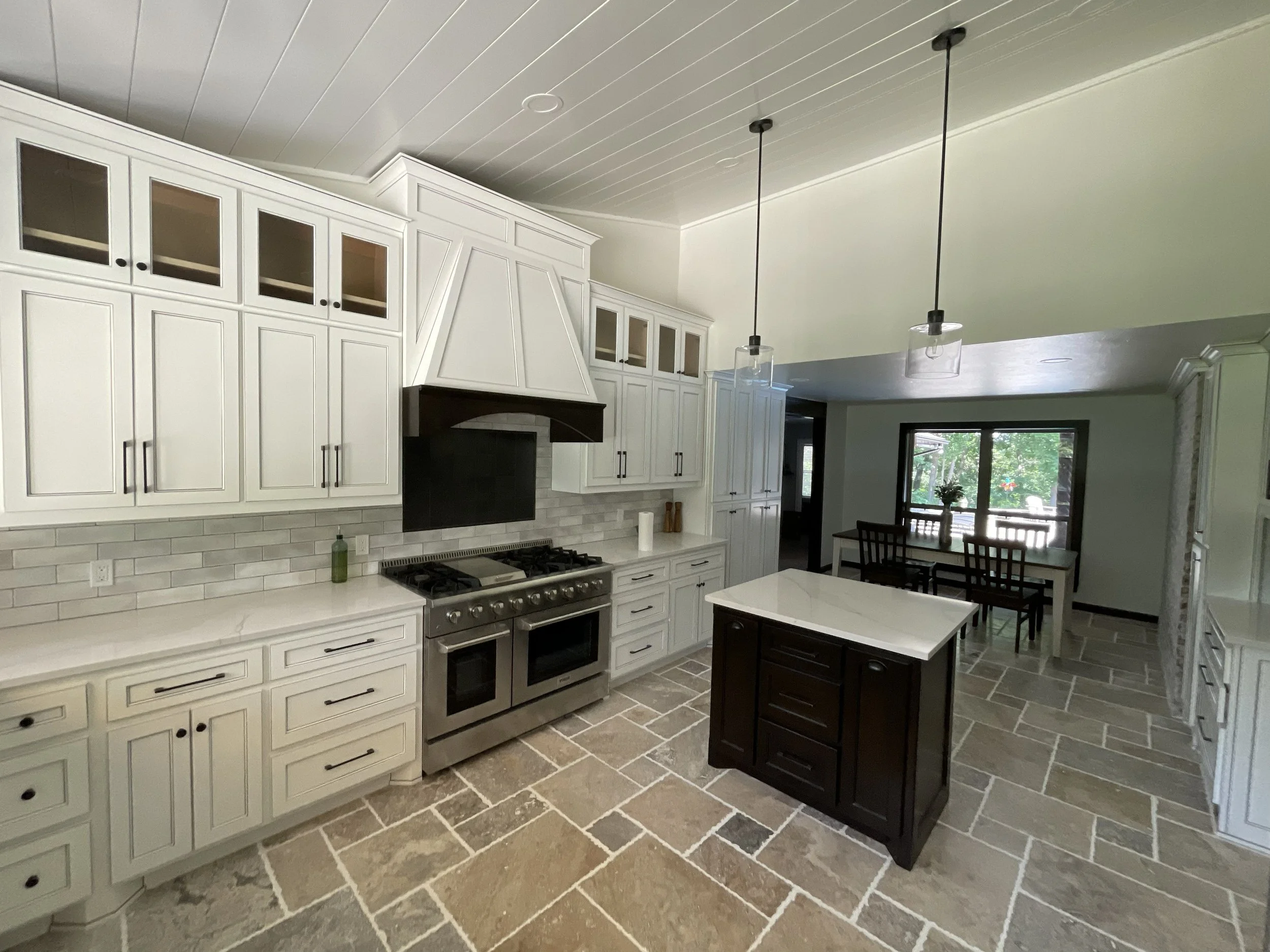 A spacious kitchen with white cabinets, a stainless steel stove, and a stone tile floor. There are high cabinets with glass doors and a black kitchen island. Large windows and a dining table are visible in the background.