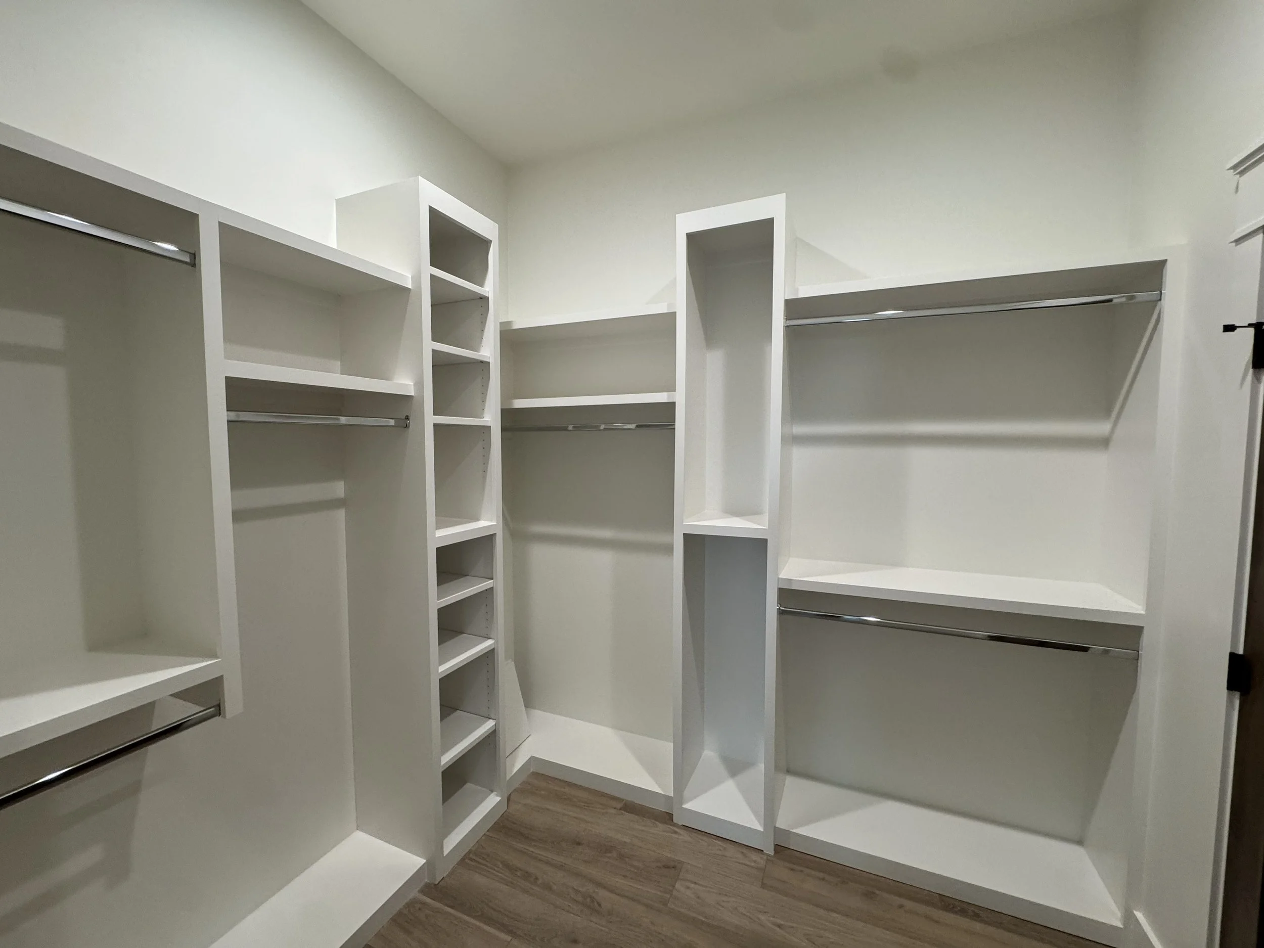 Empty walk-in closet with white built-in shelving and hanging rods, wood laminate flooring.