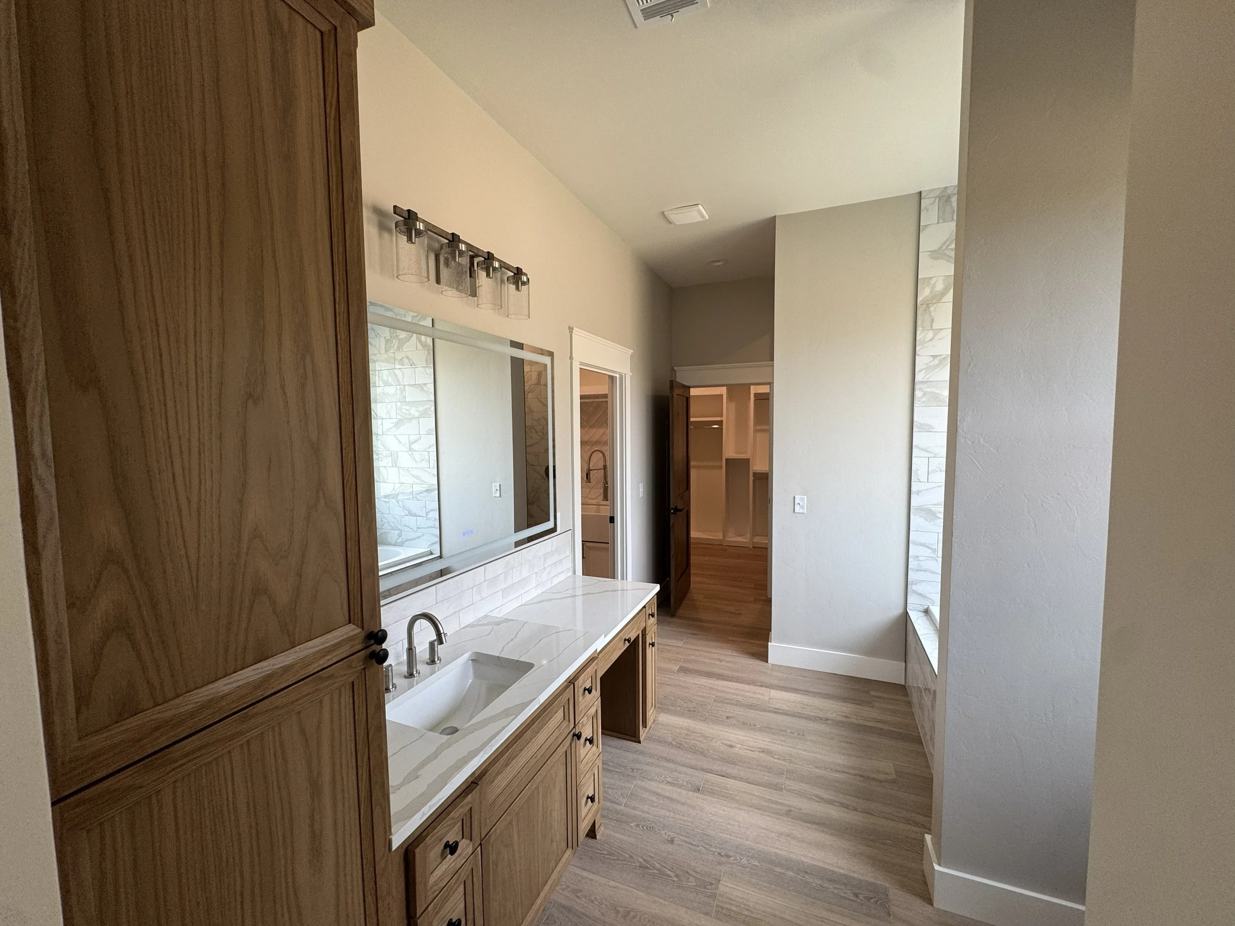 A bathroom with wooden cabinetry, a marble countertop with an undermount sink, a wall mirror, and a shower area visible through an open door.