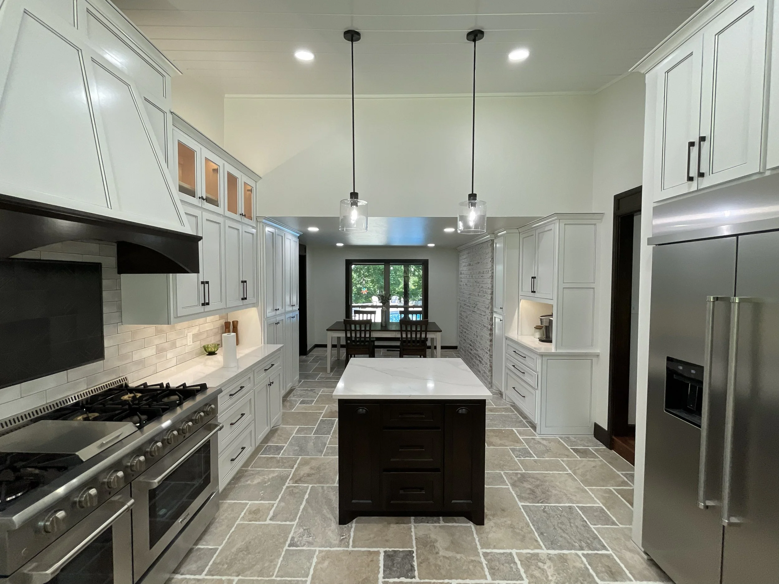 Modern kitchen with white cabinets, stainless steel appliances, two pendant lights hanging from the ceiling, a small dark island in the center, a dining area with a table and chairs in the background, large windows.