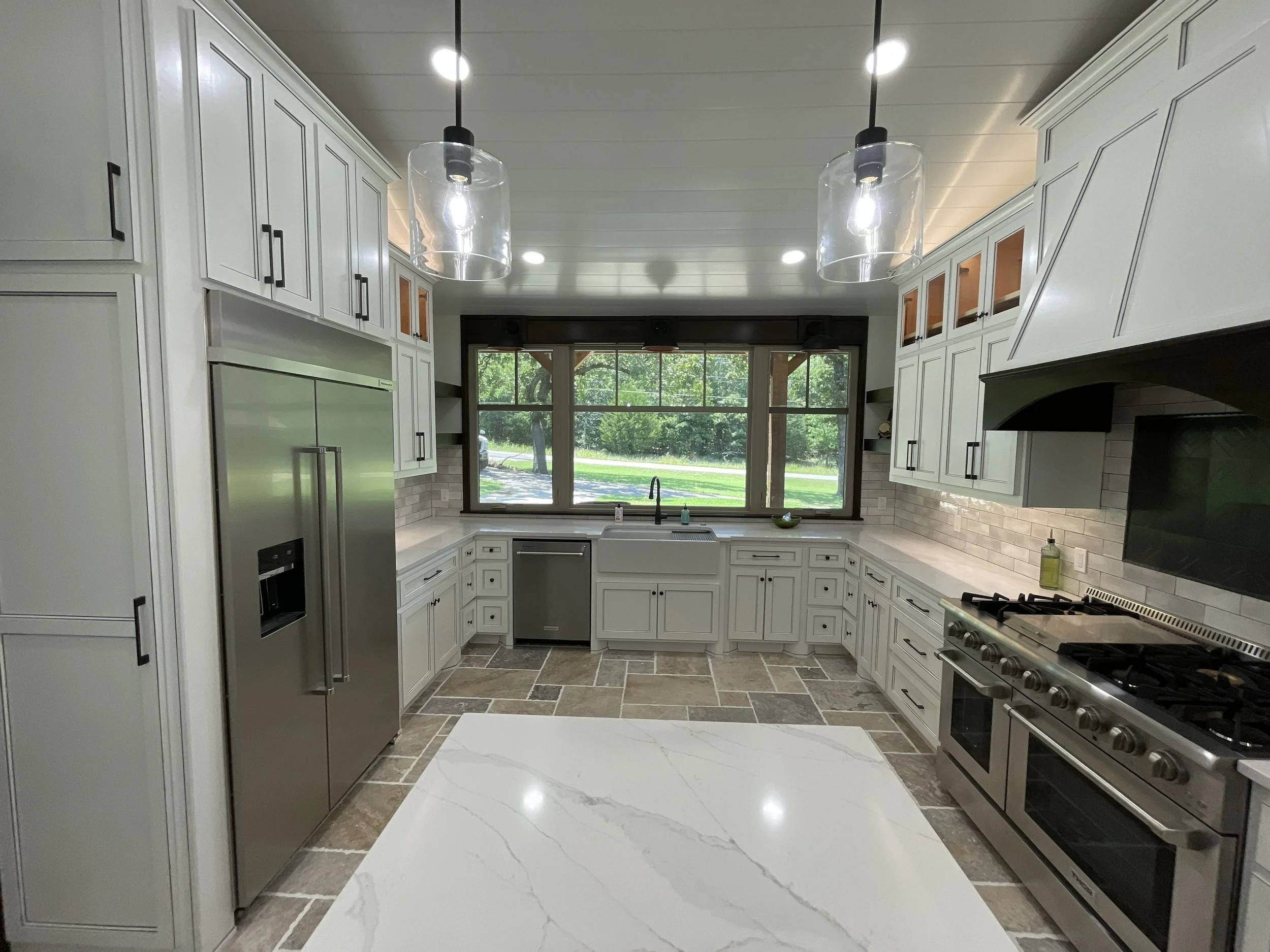 Modern kitchen with white cabinets, stainless steel appliances, large window, pendant lights, and gray tile flooring.