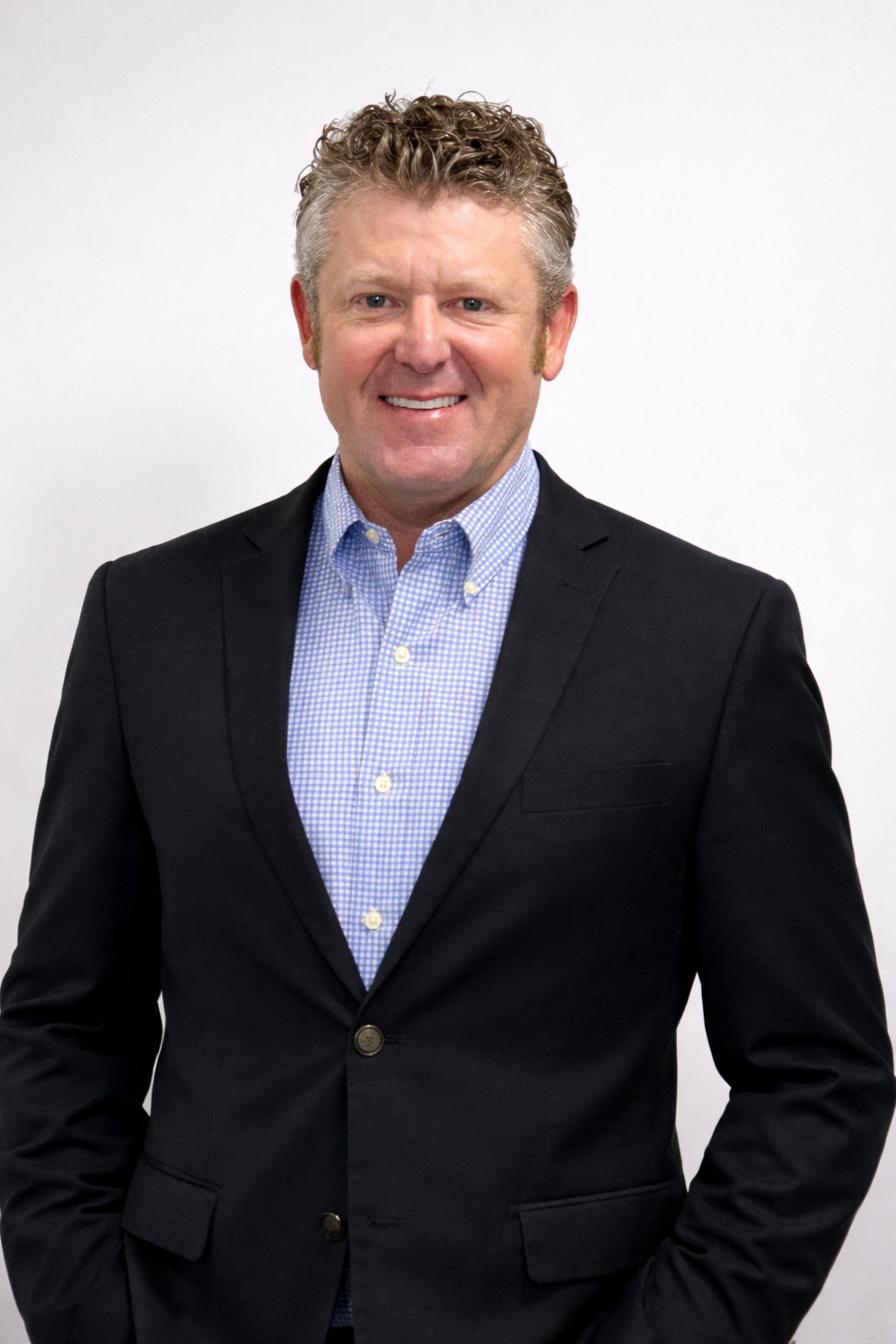 Professional portrait of a man in a black suit and light blue checkered shirt, smiling against a plain white background.