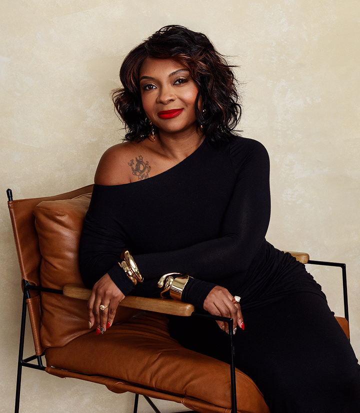A woman with dark, wavy hair and red lipstick sitting on a brown leather chair, wearing a black off-shoulder dress and gold jewelry, looking at the camera with a confident expression.