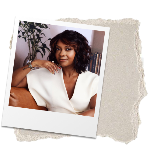 A woman with short curly dark hair sitting on a brown leather couch in a room with a potted plant and books in the background, wearing a white top.