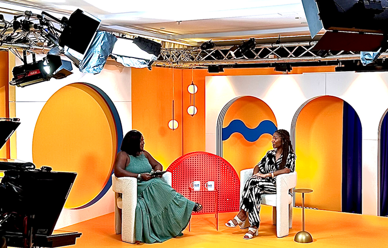 Two women sitting and talking on a colorful TV or talk show set with orange and white backdrop, circular shapes, and studio lighting.