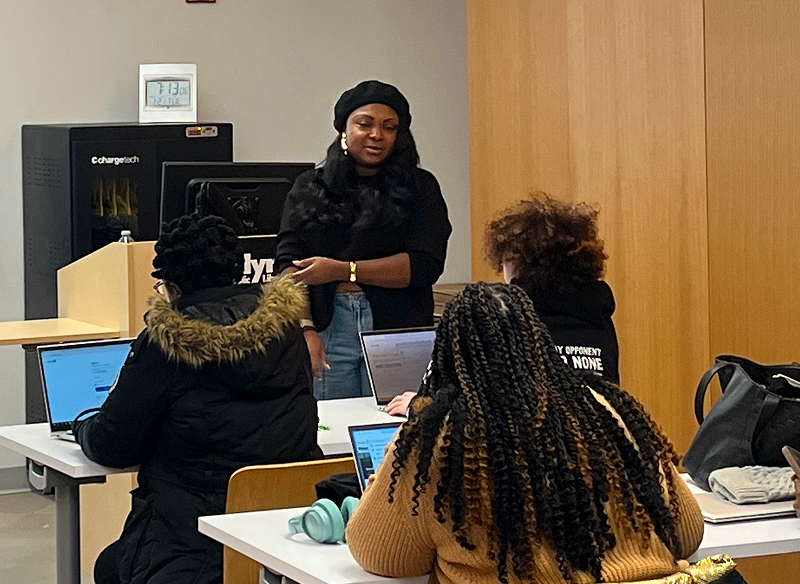 A woman standing and speaking to three women seated at tables with laptops in a classroom or meeting room.
