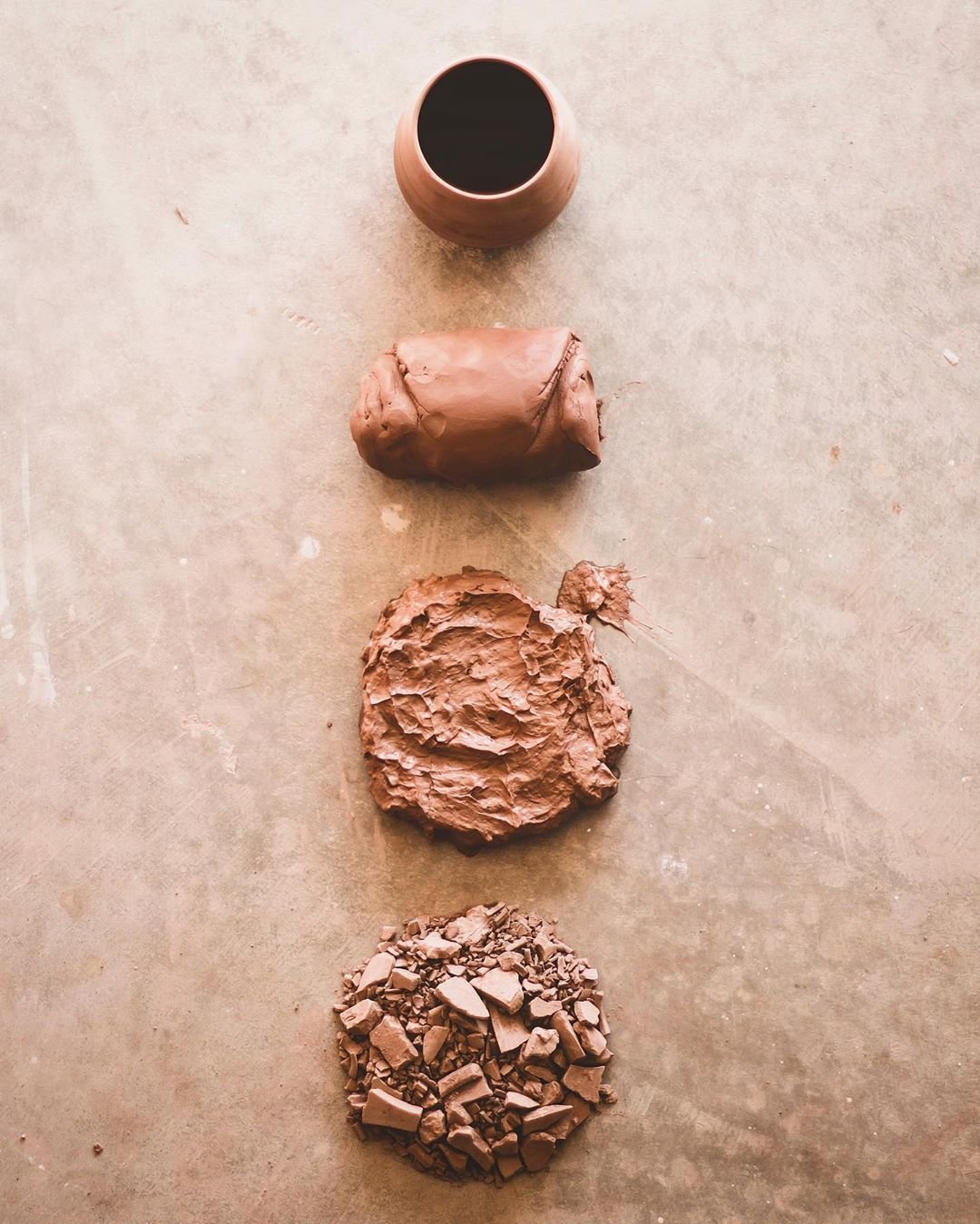 Vertical arrangement of four different types of clay: a smooth, rounded clay pot at the top filled with black liquid, then a smooth rolled piece of clay, next a rough slab of clay, and finally a pile of crushed and broken clay pieces at the bottom, on a concrete surface.