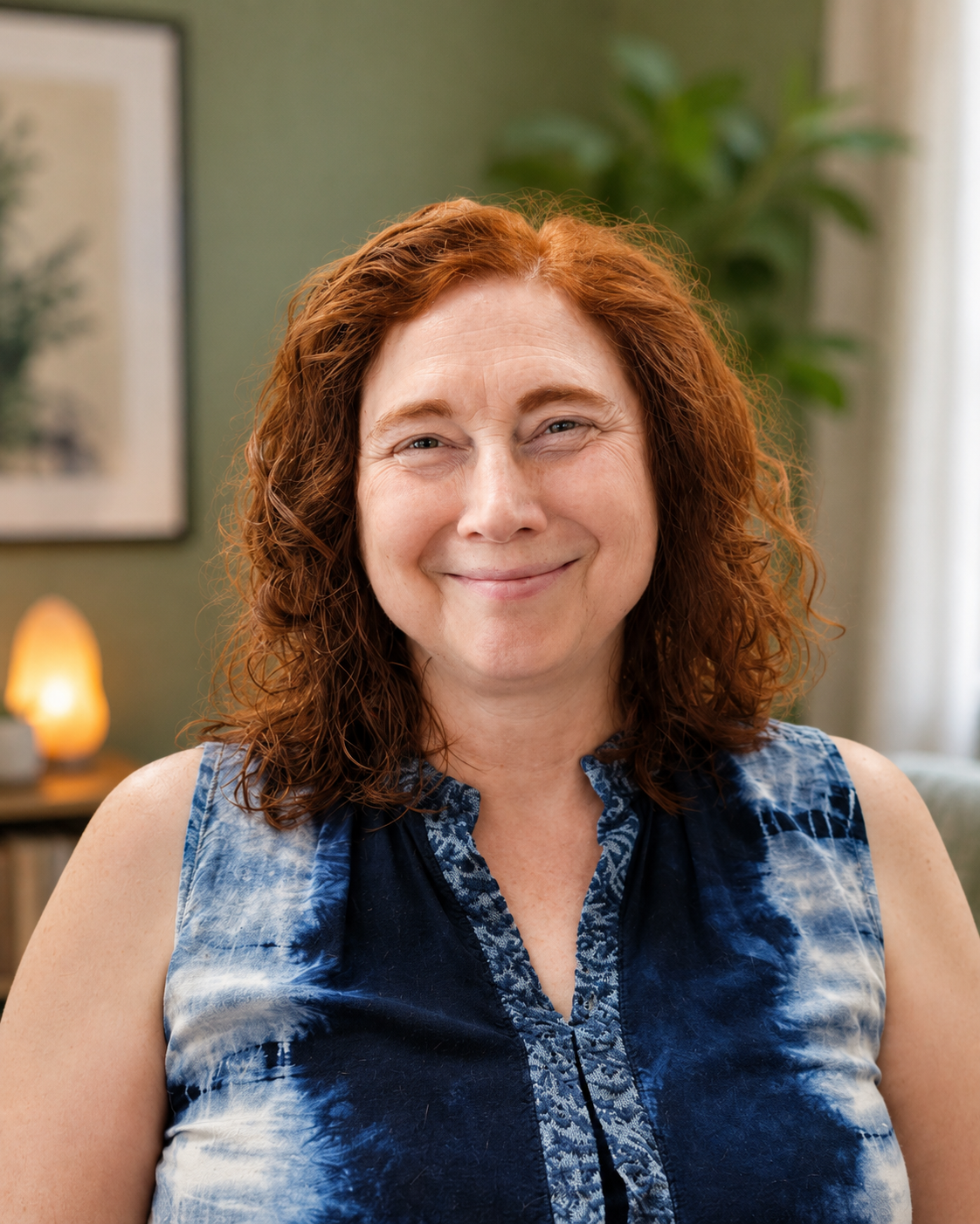 A smiling woman with curly red hair wearing a sleeveless blue tie-dye top, standing indoors with a green wall, a framed picture, a small table with a lamp, and a plant in the background.