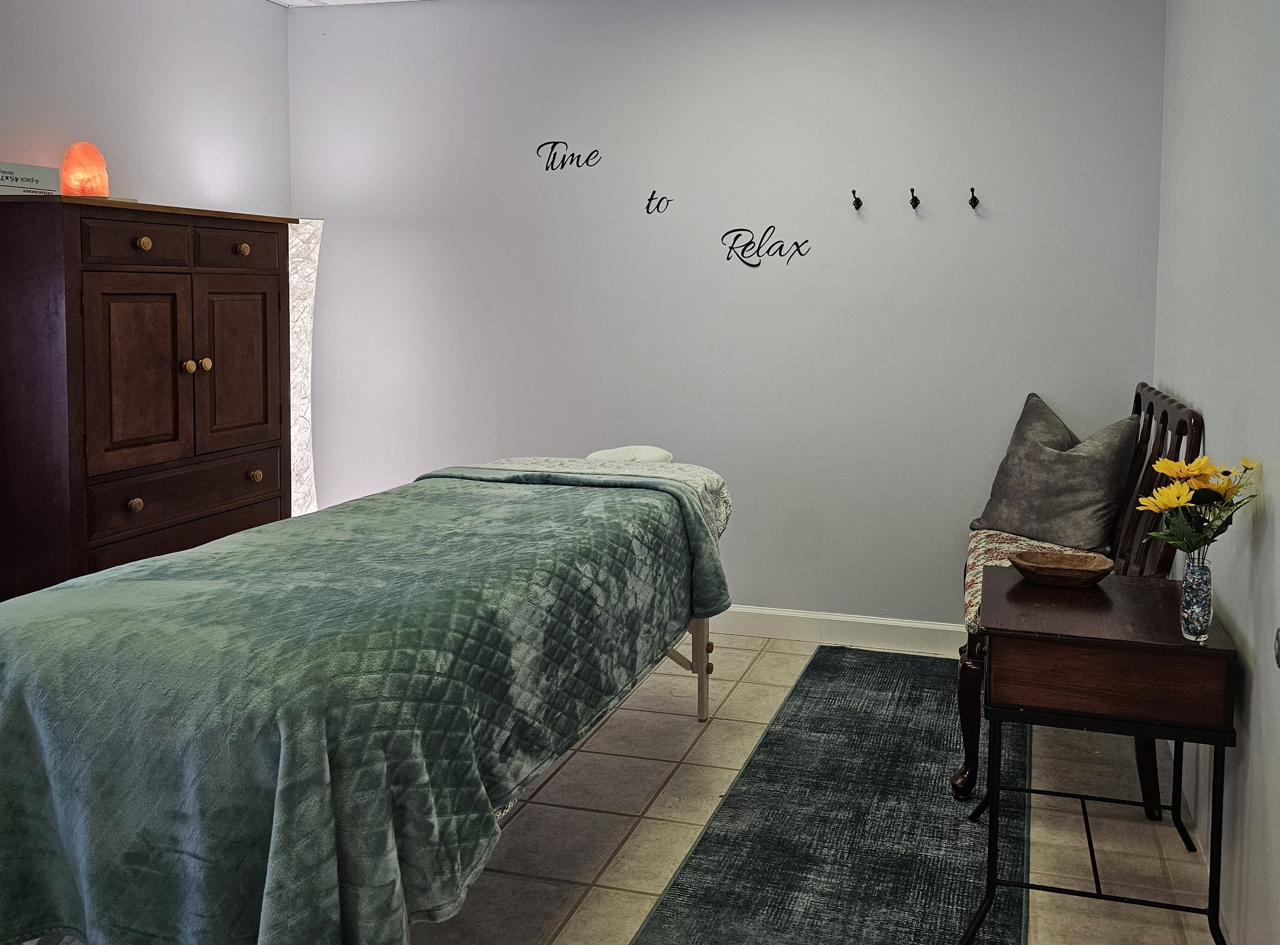 A cozy room with a bed covered in a green quilt, a wooden dresser with rounded knobs, a wooden side table with a vase of yellow flowers, and a wall decal that reads 'Time to Relax'.
