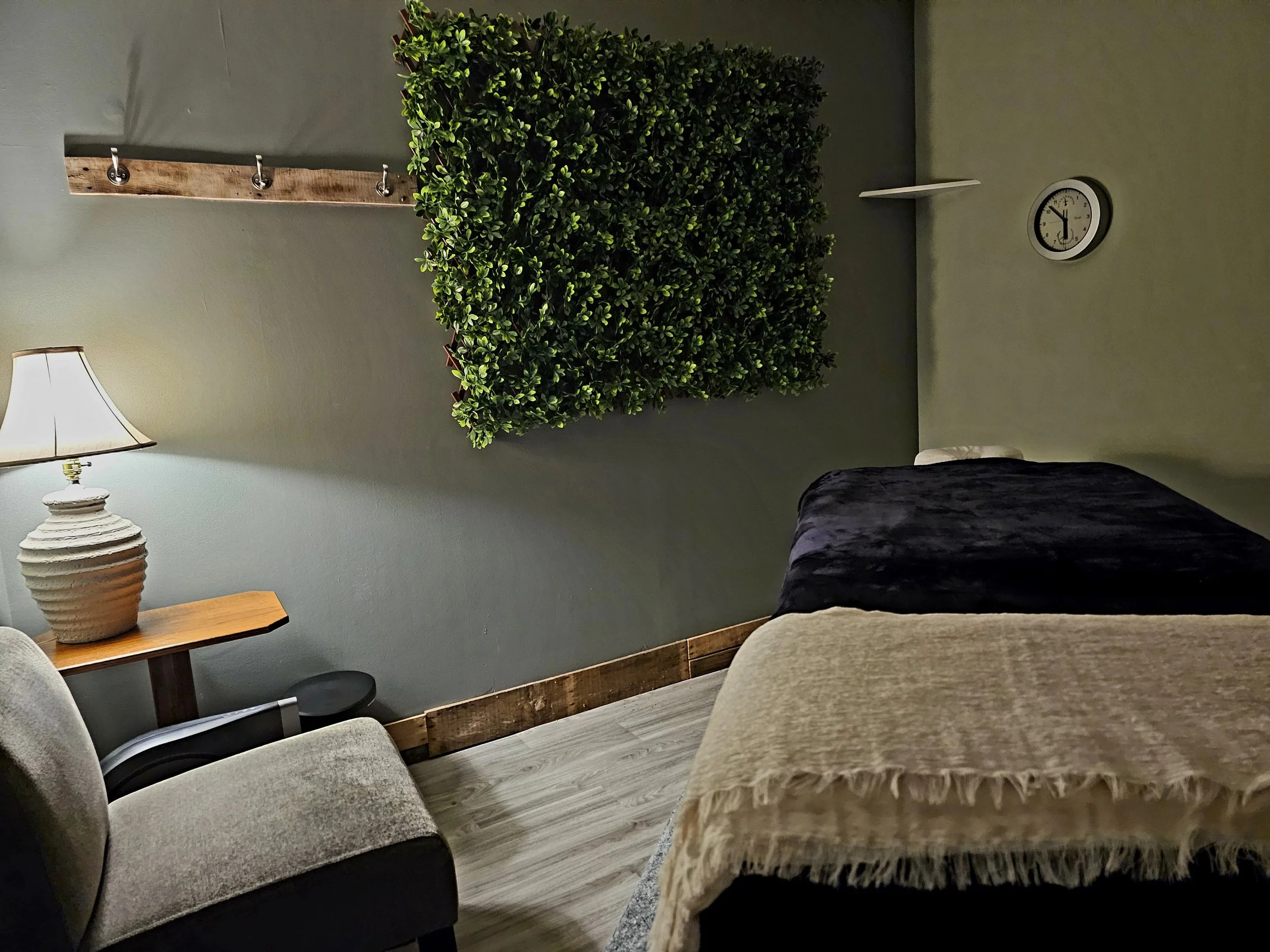 Living room with a green wall, a hanging plant, a white clock, a side table with a lamp, and upholstered chairs.