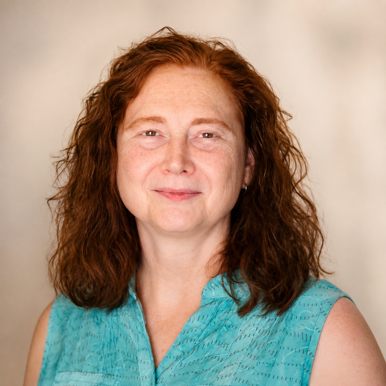 A woman with curly red hair wearing a sleeveless blue top standing against a plain beige background.