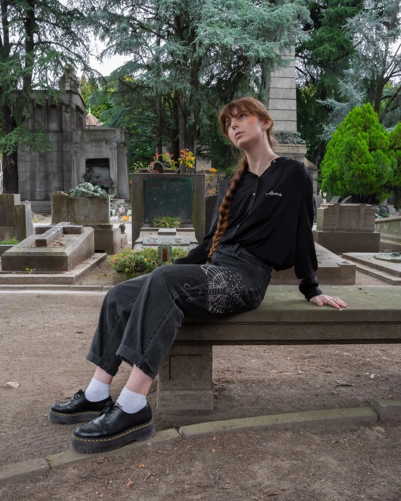 A young woman with red hair in a braid sits on a stone bench in a cemetery, surrounded by tombstones, trees, and greenery, looking off to the side.