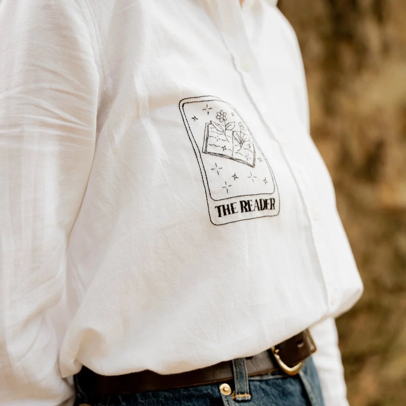 Close-up of a white shirt with a printed design of a book, flowers, and stars, labeled "The Reader."