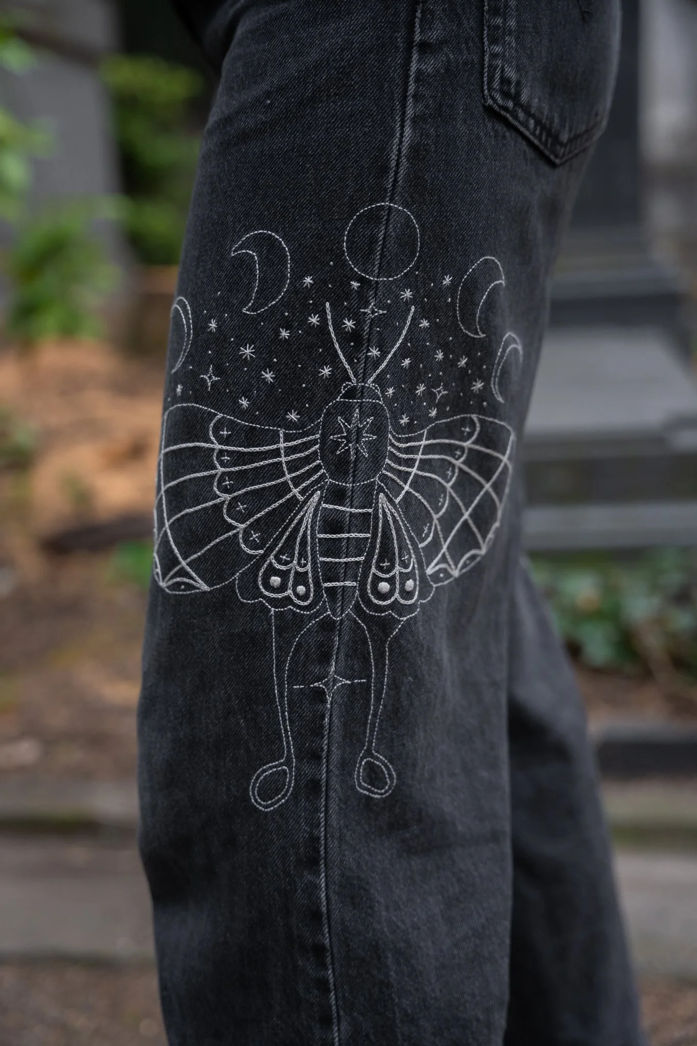 Close-up of black denim jeans with white line art design of a butterfly, moon phases, and stars printed on the leg.