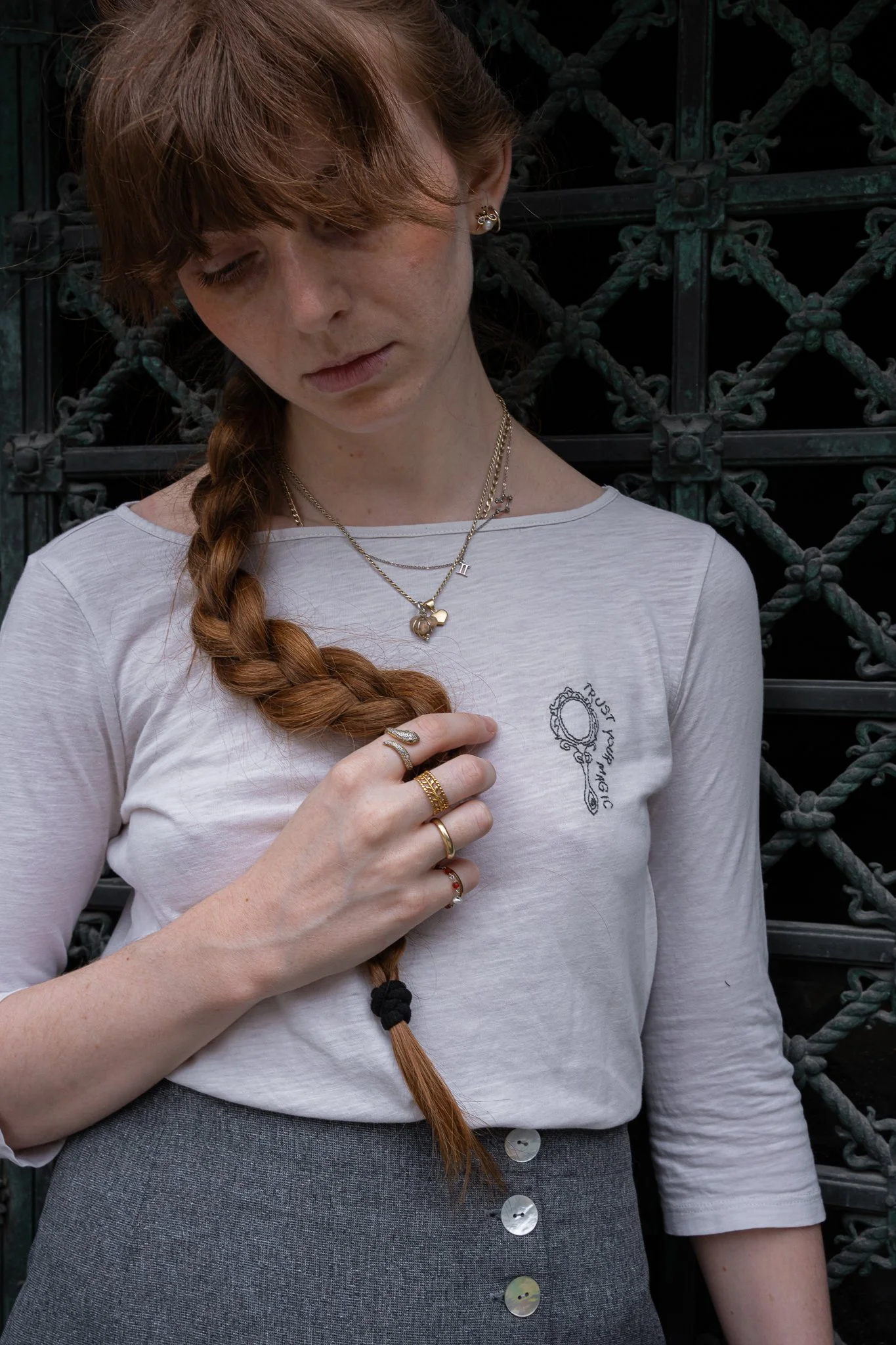 A woman with light skin and long, red hair in a braid wearing a white t-shirt and gray skirt, standing against a decorative black metal fence. She is looking down and is adorned with multiple gold necklaces and rings.