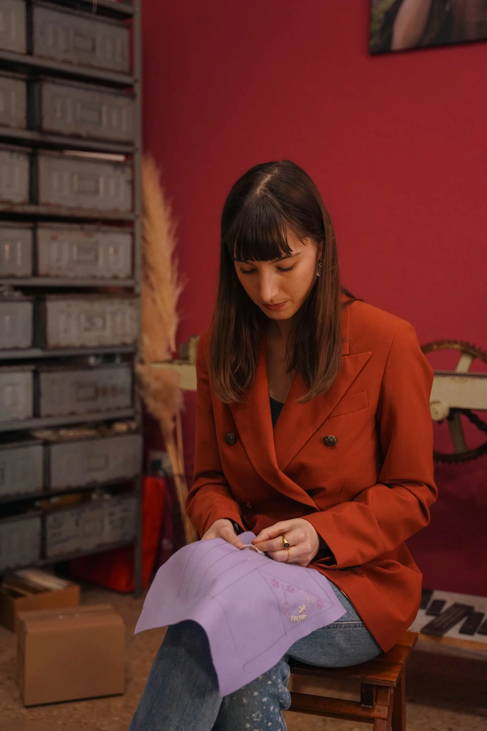 Una donna con capelli castani indossa una giacca rossa e sta ricamando seduta su uno sgabello. Lo sfondo mostra pareti rosse, scaffali con scatole e vari oggetti decorativi.