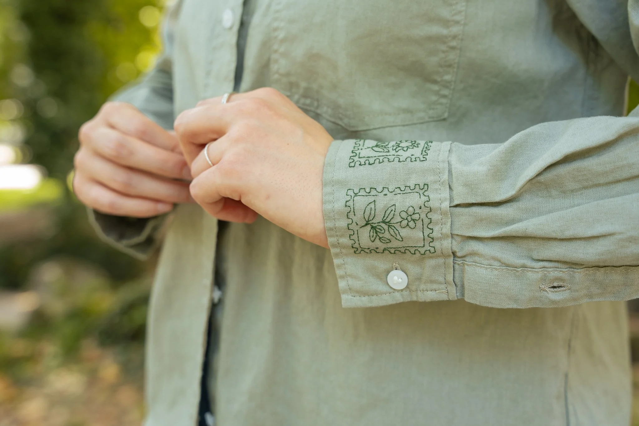 Close-up of a person’s hands clasped together, wearing a sage green shirt with  dark green embroidered designs on the sleeve, outdoors with blurred greenery in the background.