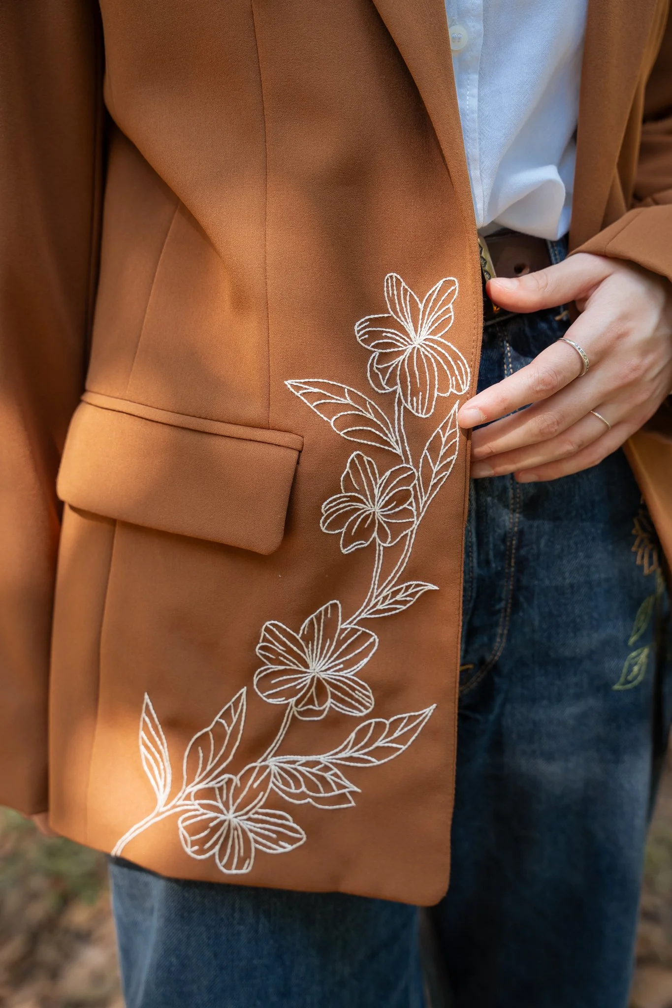 Close-up of a person wearing a brown blazer with white floral embroidery along the edge, white shirt, and blue jeans.