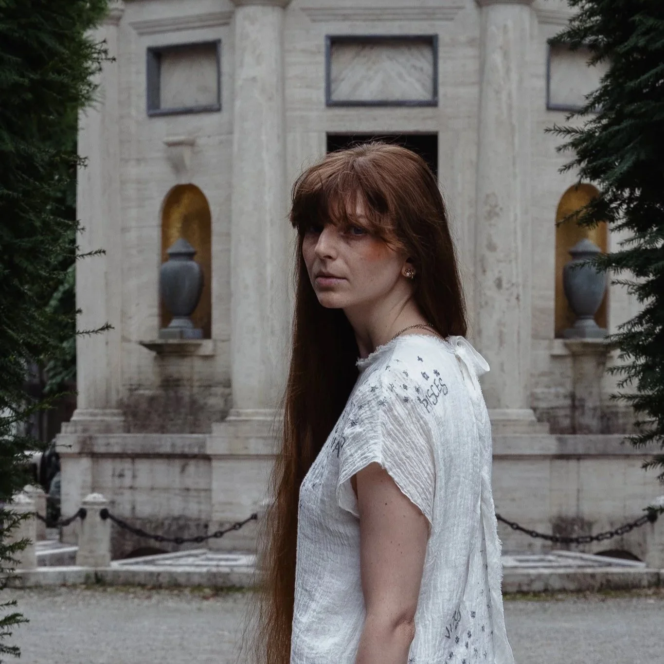 A woman with long red hair and a white blouse stands outdoors with an ancient stone building featuring columns, arches, and urn sculptures in the background, framed by green trees.