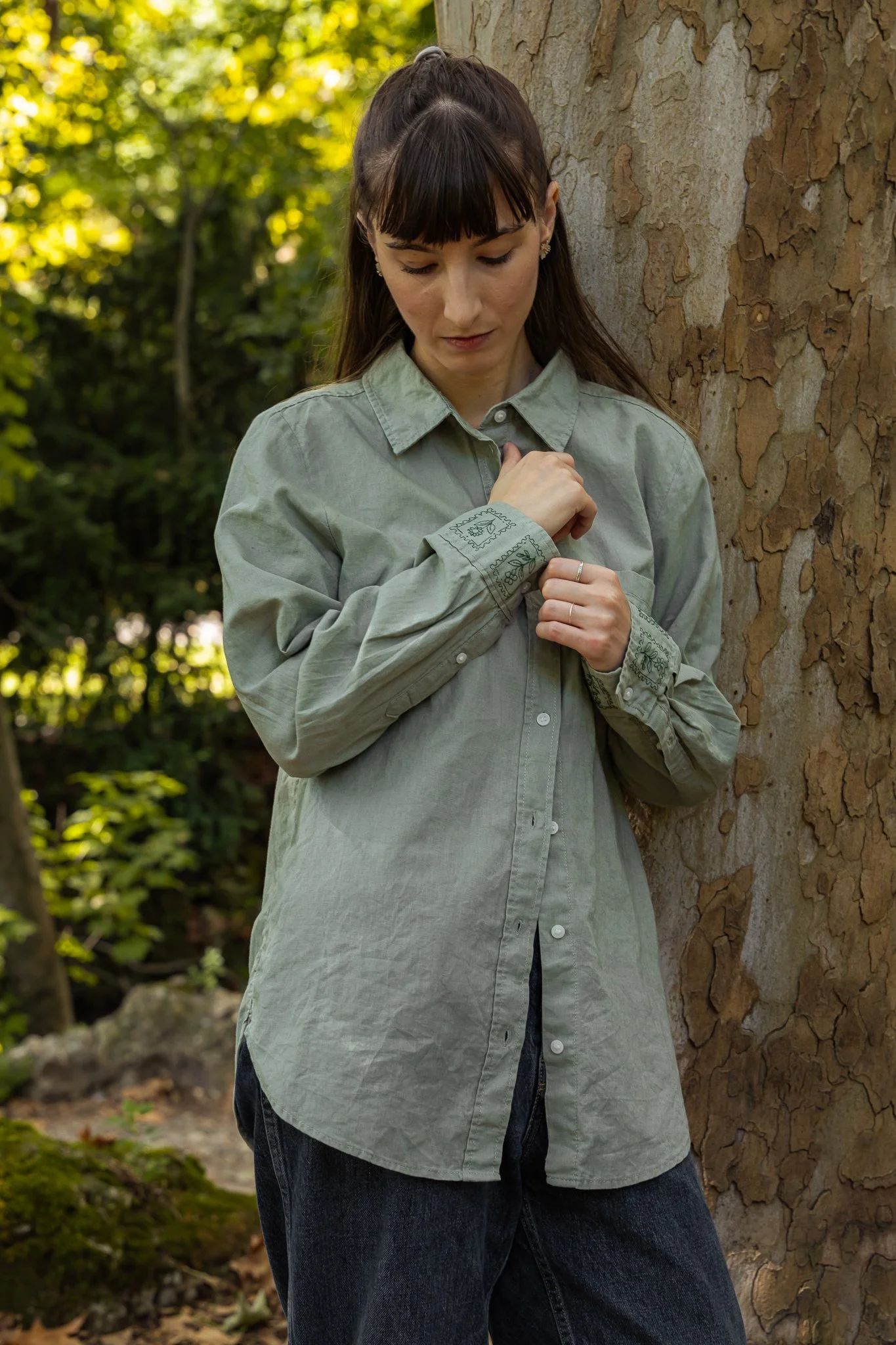 A young woman with dark hair in a ponytail, wearing earrings, a loose light green button-up shirt with embroidered cuffs, and dark jeans, standing outdoors next to a tree with textured bark. She has her eyes closed and is touching her shirt near her 