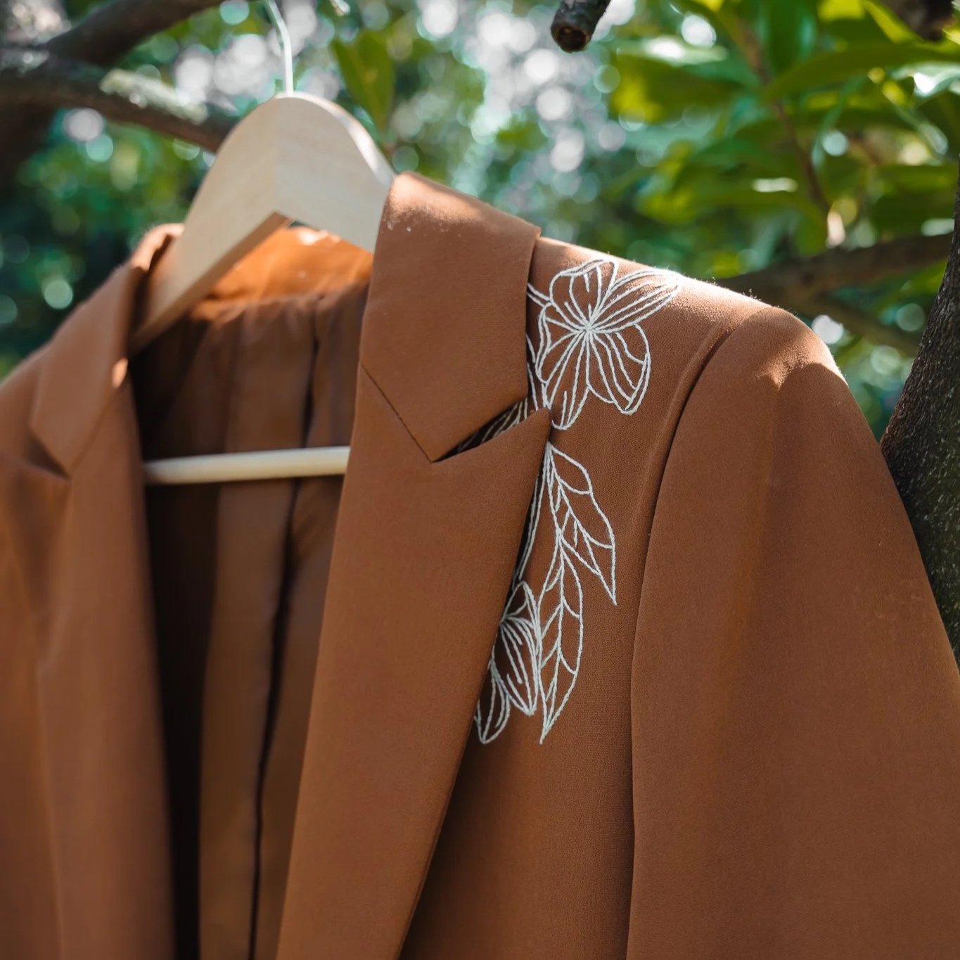 A brown blazer jacket hanging on a wooden hanger outdoors on a tree branch, with white floral embroidery near the collar, and green leaves in the background.