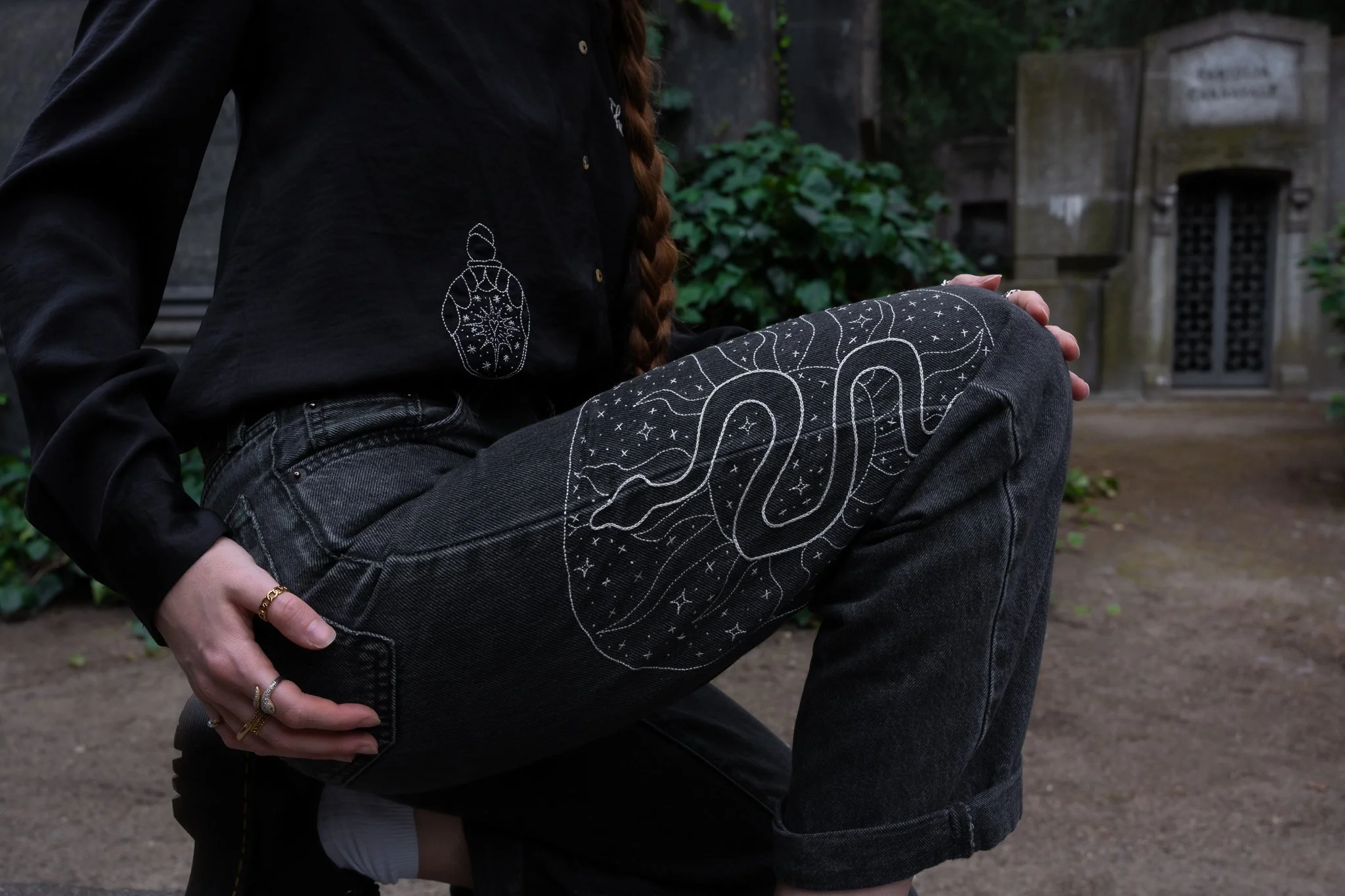 A person sitting outdoors, wearing a black shirt with embroidered designs and gray jeans with celestial patterns, cradling their knee while resting, with a stone structure and greenery in the background.