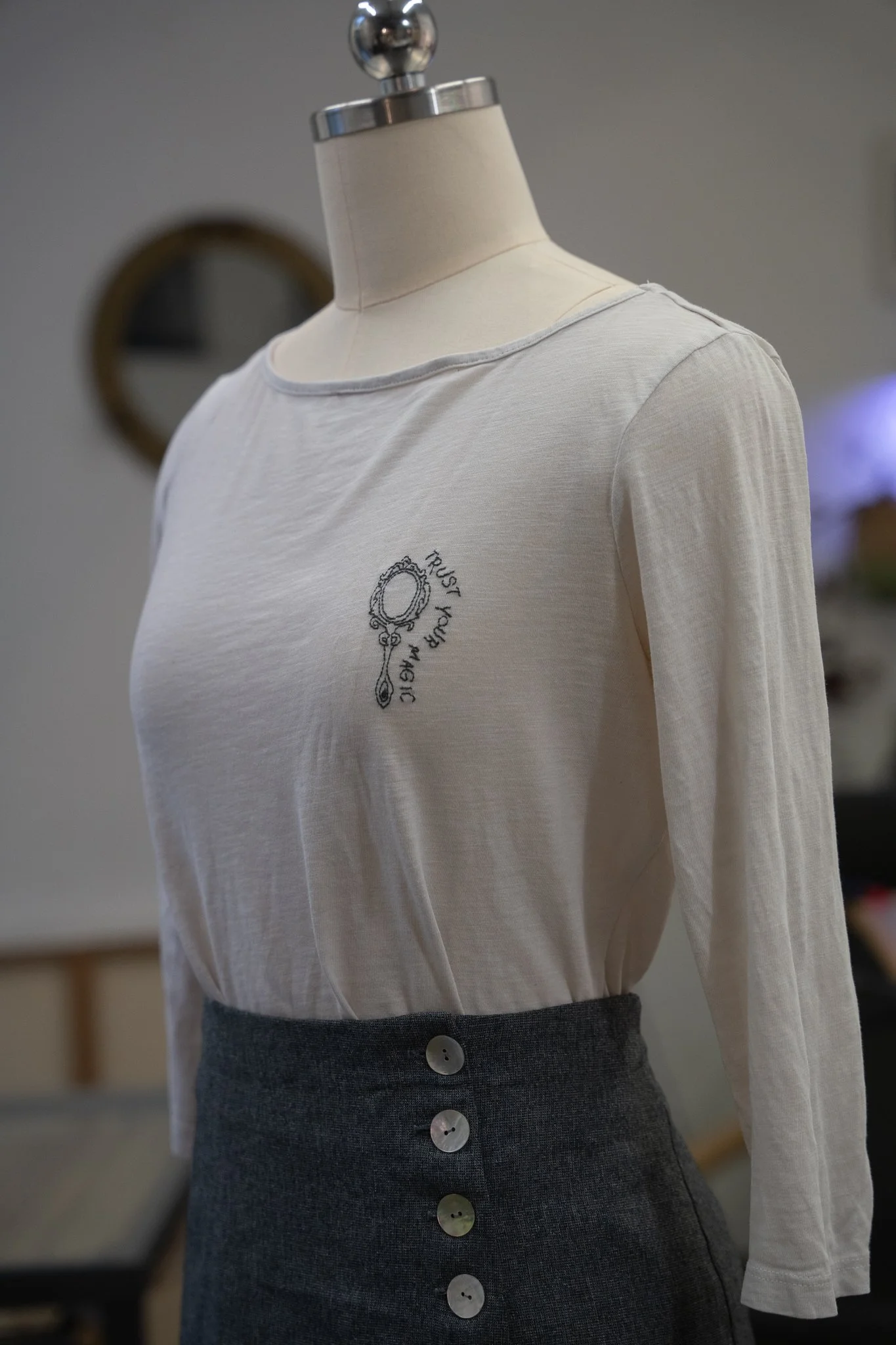 A mannequin dressed in a white long-sleeve shirt with a small embroidery on the left side and a gray high-waisted skirt with large buttons.