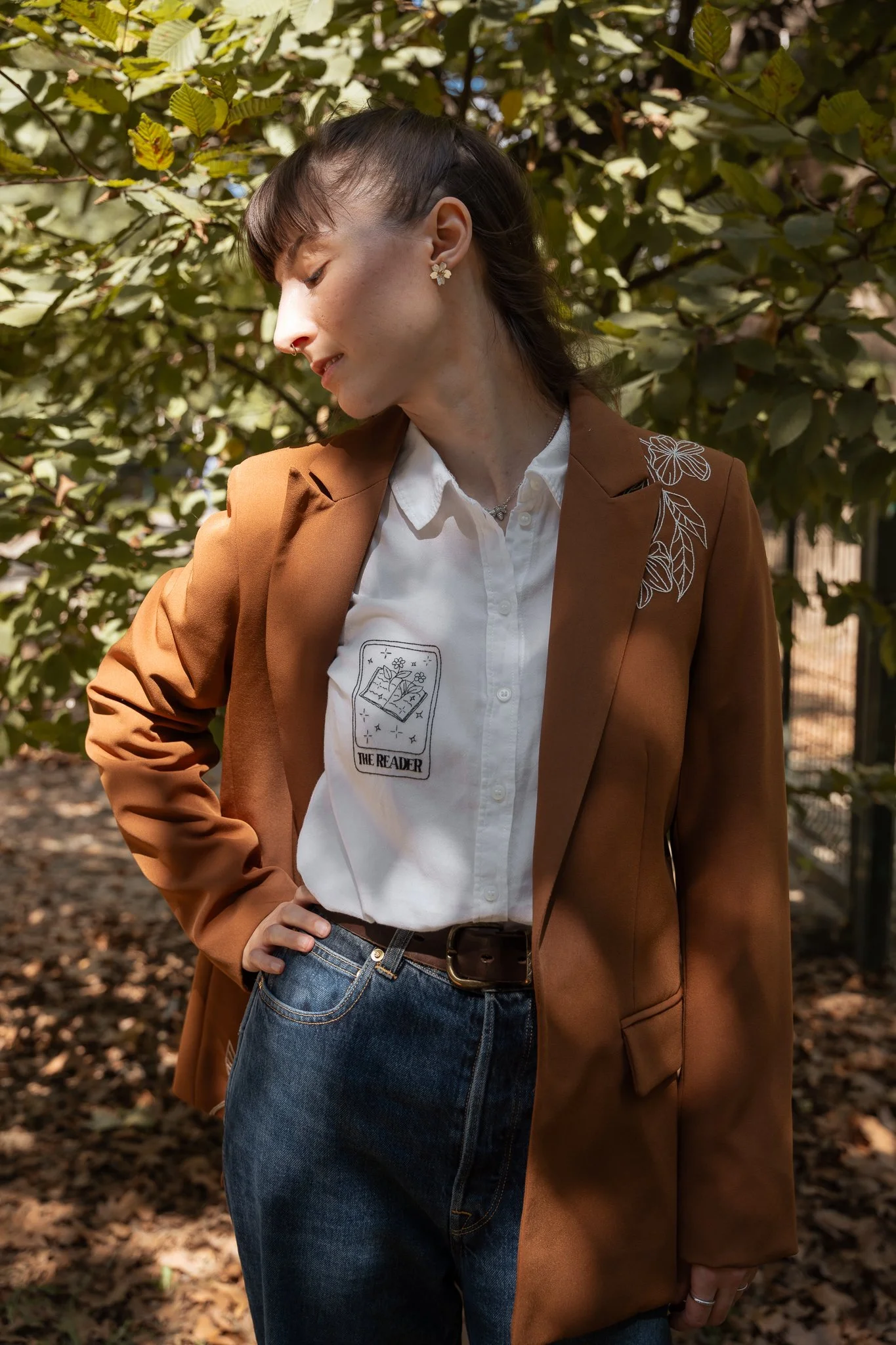 A woman with dark hair in a ponytail, wearing a white shirt with a printed design, a brown blazer with floral embroidery, and blue jeans, standing outdoors among green and brown foliage, with her eyes closed and one hand on her hip.